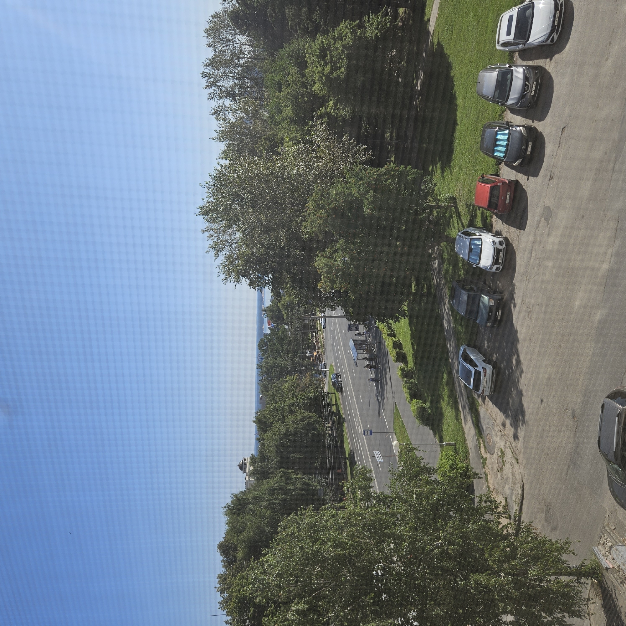 The image depicts a street scene with a clear blue sky. In the foreground, a row of parked cars are visible on a paved area. From left to right, these include a dark SUV, a white sedan, a dark green SUV, a white SUV, a red hatchback, a dark gray SUV, a silver sedan, and a white sedan. Behind the cars, a street runs horizontally across the image, with a bus stop on the left side and trees lining the opposite side. A few pedestrians are present near the bus stop. In the background, a body of water can be seen, with trees and buildings on the horizon. A domed building is prominent among the structures. The environment appears to be a suburban or semi-urban area in Cheboksary, Russia.