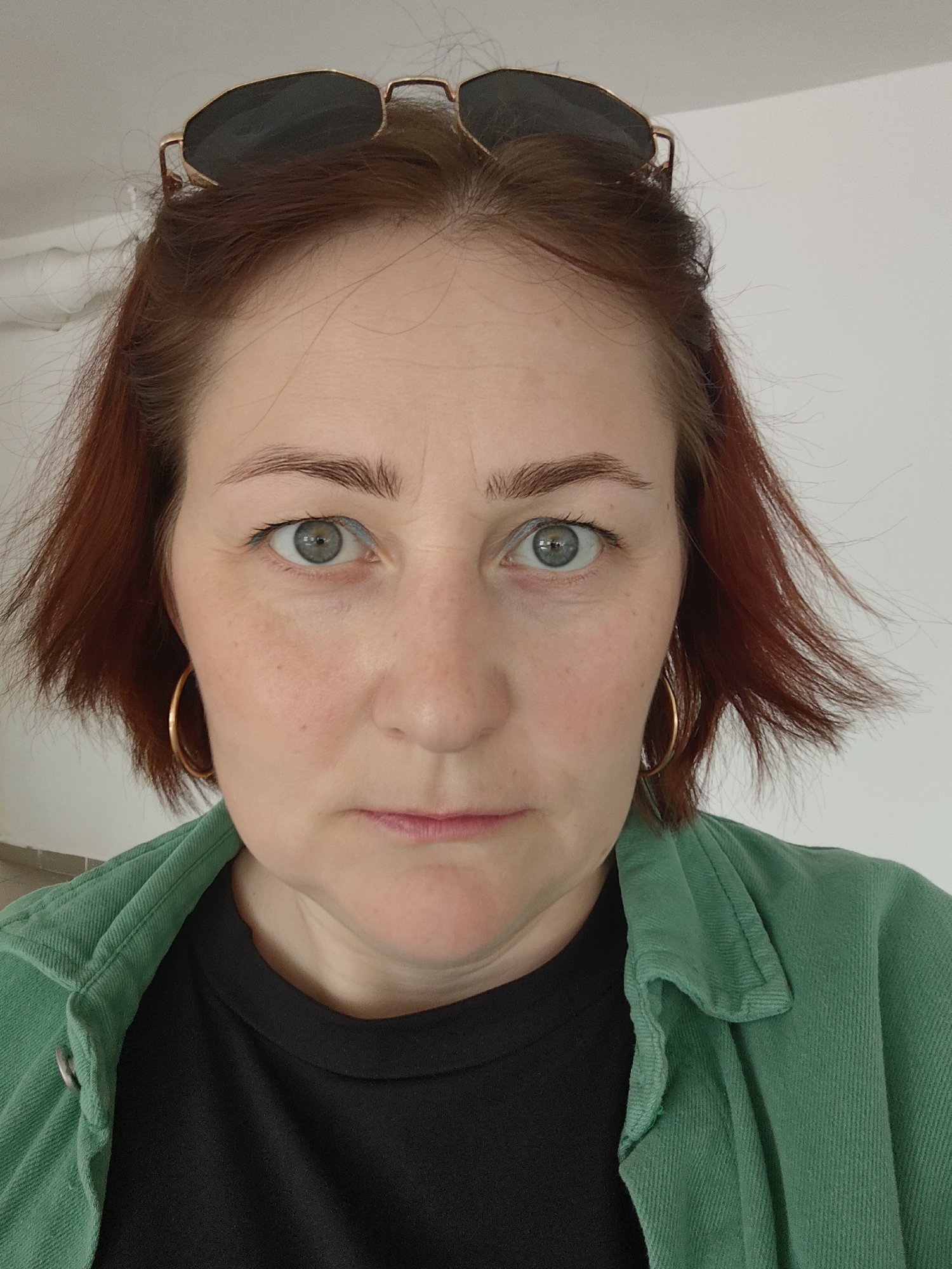 A close-up, slightly angled portrait shows a woman with fair skin, light eyes, and short, auburn hair. Sunglasses are perched on top of her head. She is wearing gold hoop earrings, a black t-shirt, and a green corduroy shirt or jacket. The background is a plain white wall. There is no indication of the location being Марковский, Russia.