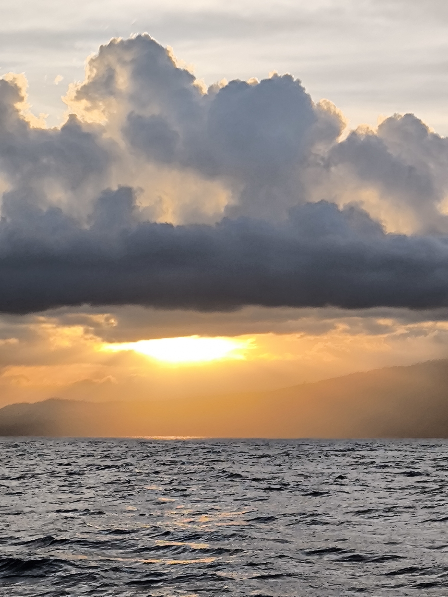The image depicts a sunset over water, likely a sea or large lake. The location is Unknown, Indonesia, though this is only asserted and not verifiable from the image itself. The sky is dominated by large, cumulonimbus clouds, some of which are dark grey, while others are illuminated with a golden hue from the setting sun. A bright, horizontal band of sunlight breaks through a lower layer of clouds, casting a strong glare. The sun's rays reflect off the water's surface, creating a shimmering effect. In the distance, silhouettes of landmasses, possibly islands or mountains, are visible. The water is choppy, with small waves creating texture and reflecting the light.