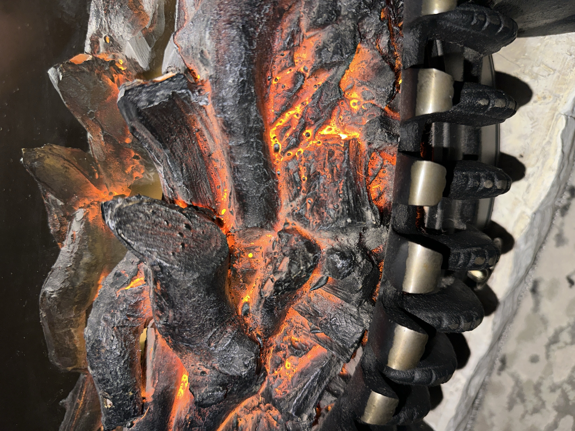 The image shows a close-up of an artificial fireplace with faux logs and glowing embers. The logs are dark gray and black, with textured surfaces that mimic burnt wood. Between the logs, bright orange and yellow light emanates, simulating the glow of embers. The logs are arranged in a heap, suggesting a crackling fire. Below the logs is a metal grate, also black, with metallic accents that appear to hold the logs in place. The background is dark, and the foreground shows a mottled, light-colored surface, possibly a hearth or floor. The scene is static and does not depict any activity or individuals. The location is Unknown, Unknown.