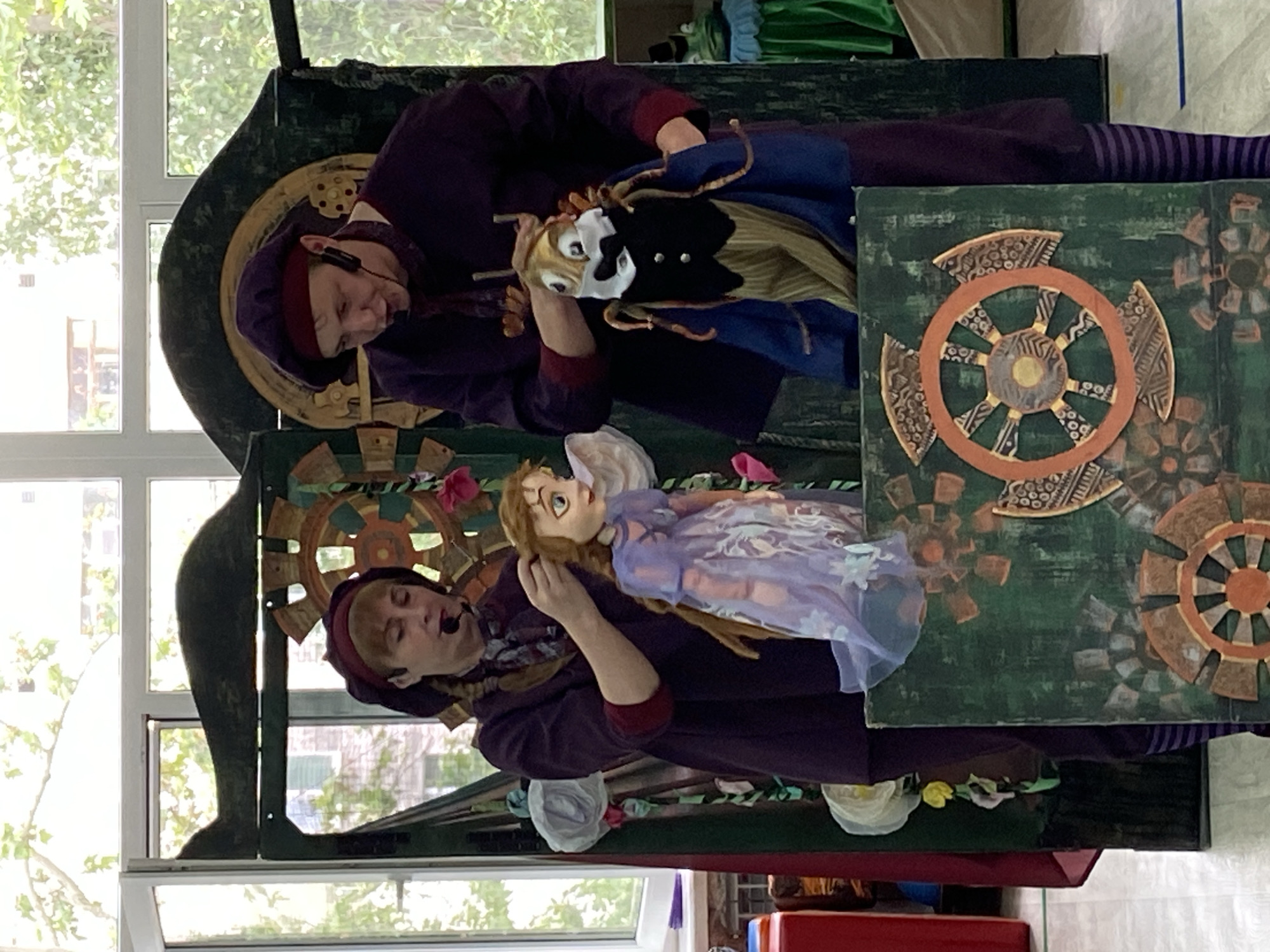 Two individuals, dressed in purple costumes and maroon berets, are operating puppets in front of a stage-like backdrop. The backdrop is decorated with green painted panels featuring a large, intricate circular design resembling a gear or a wheel. The individual on the left is manipulating a doll dressed in a light purple dress, while the individual on the right is operating a puppet that resembles an insect with a suit and tie. Both individuals appear to be speaking or performing, as indicated by their open mouths and microphones attached to their ears. The setting appears to be indoors, with large windows in the background that let in natural light and show greenery outside. There are also some decorative elements on the stage, such as paper flowers. The floor is light-colored and reflective.