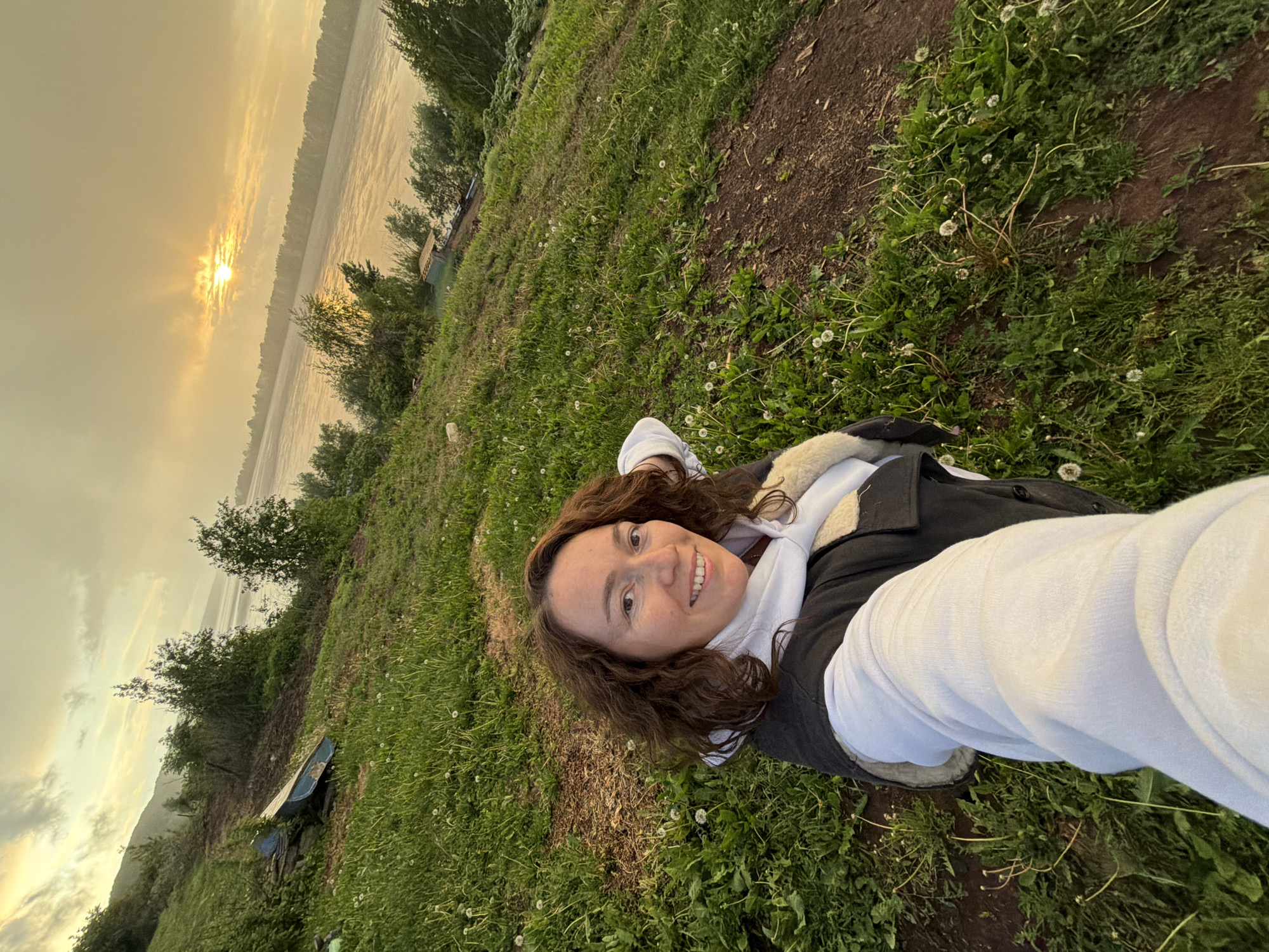 A smiling woman with brown, wavy hair is taking a selfie, appearing to be lying or reclining on a grassy bank. She is wearing a white long-sleeved top under a dark, possibly fleece-lined, vest or jacket.
The scene is an outdoor setting beside a wide body of water, likely a river, with green vegetation along the banks. The image captures either a sunrise or sunset, with the sun low on the horizon, casting a bright, golden light that reflects on the water and illuminates the sky with warm, hazy hues. In the background, across the water, a forested or tree-lined bank is visible. Closer to the foreground, on the grassy bank where the woman is, there are scattered white dandelions and patches of exposed dirt. A small, blue boat is partially visible on the riverbank. The woman's joyful expression suggests a pleasant and serene moment in this natural environment, likely in Ольховка, Russia. No visible text is present.