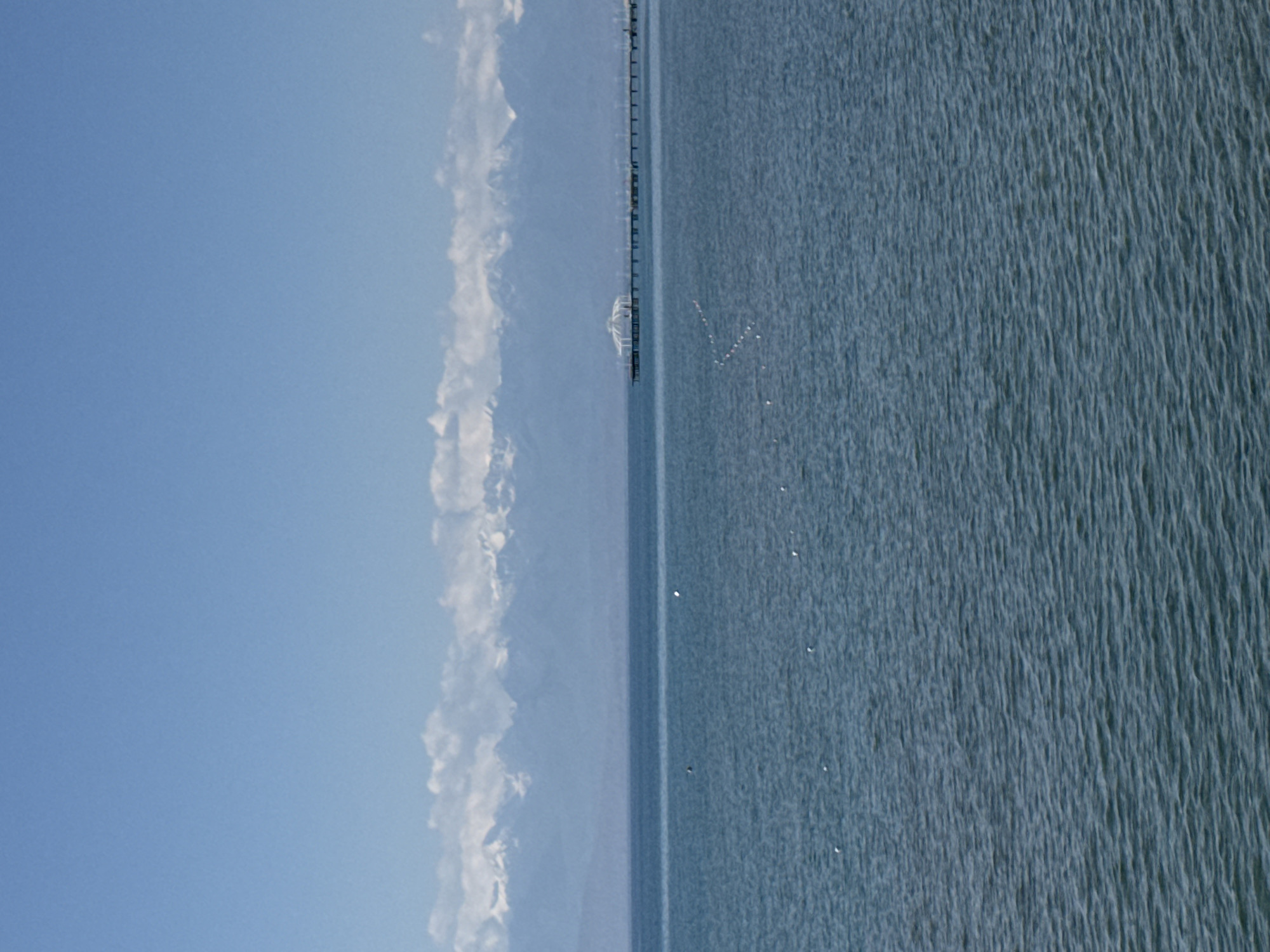 A wide shot captures a serene, vast body of blue water under a clear, pale blue sky. In the distance, a long, narrow pier extends from the horizon, featuring a white, domed structure at its end. Beyond the pier and water, a range of snow-capped mountains forms a majestic backdrop, their peaks softened by a thin layer of white clouds. The water's surface is gently rippled, and a few small, white objects, possibly buoys or birds, are scattered across it. The scene suggests a tranquil lakeside or coastal setting in Koshkol', Kyrgyzstan, likely during the daytime.