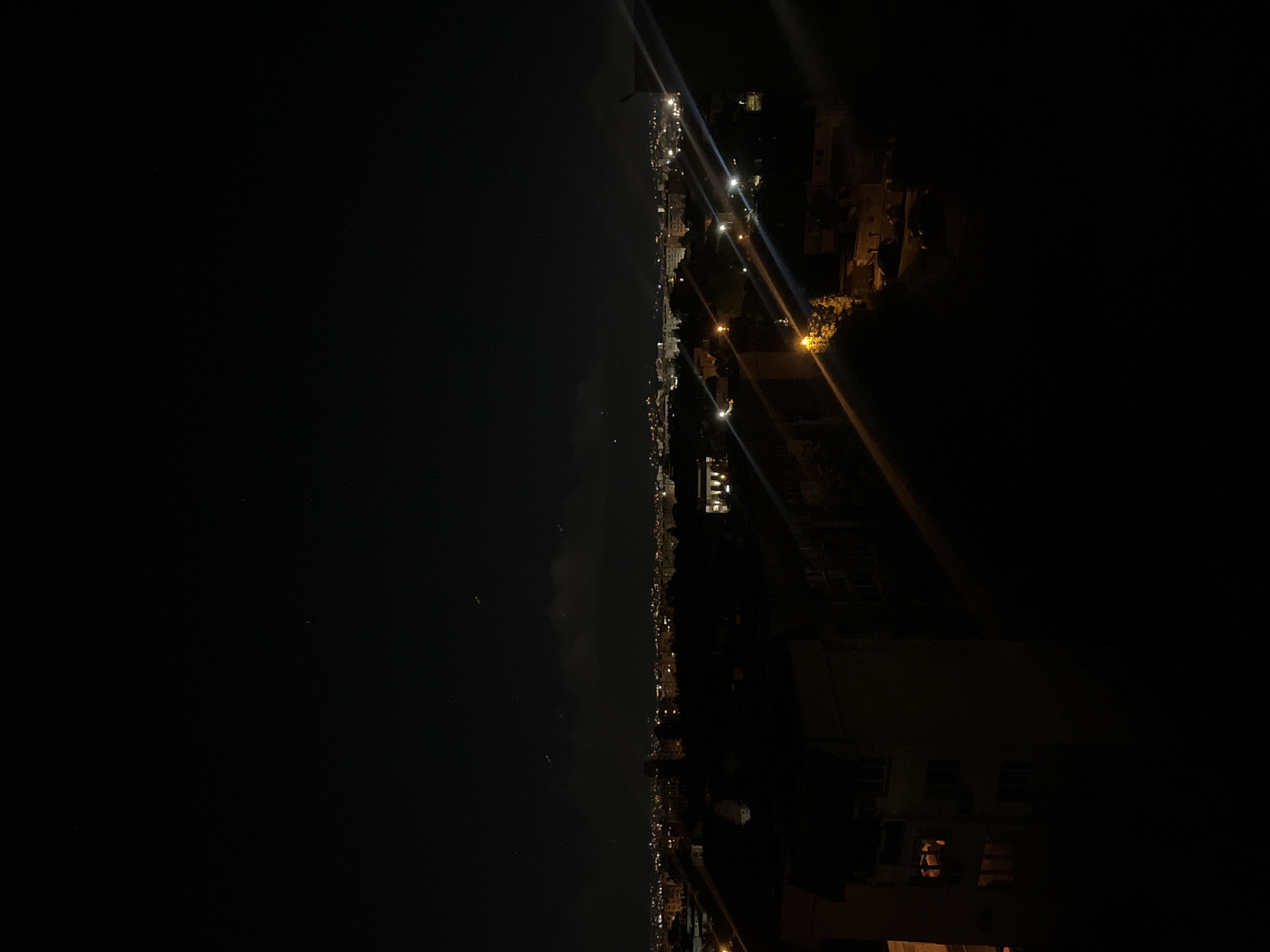 The image depicts a night scene in Sofia, Bulgaria. A dark sky and sea are on the left. A line of lights suggests a coastline. Buildings with illuminated windows are on the right, with streetlights casting rays into the darkness.