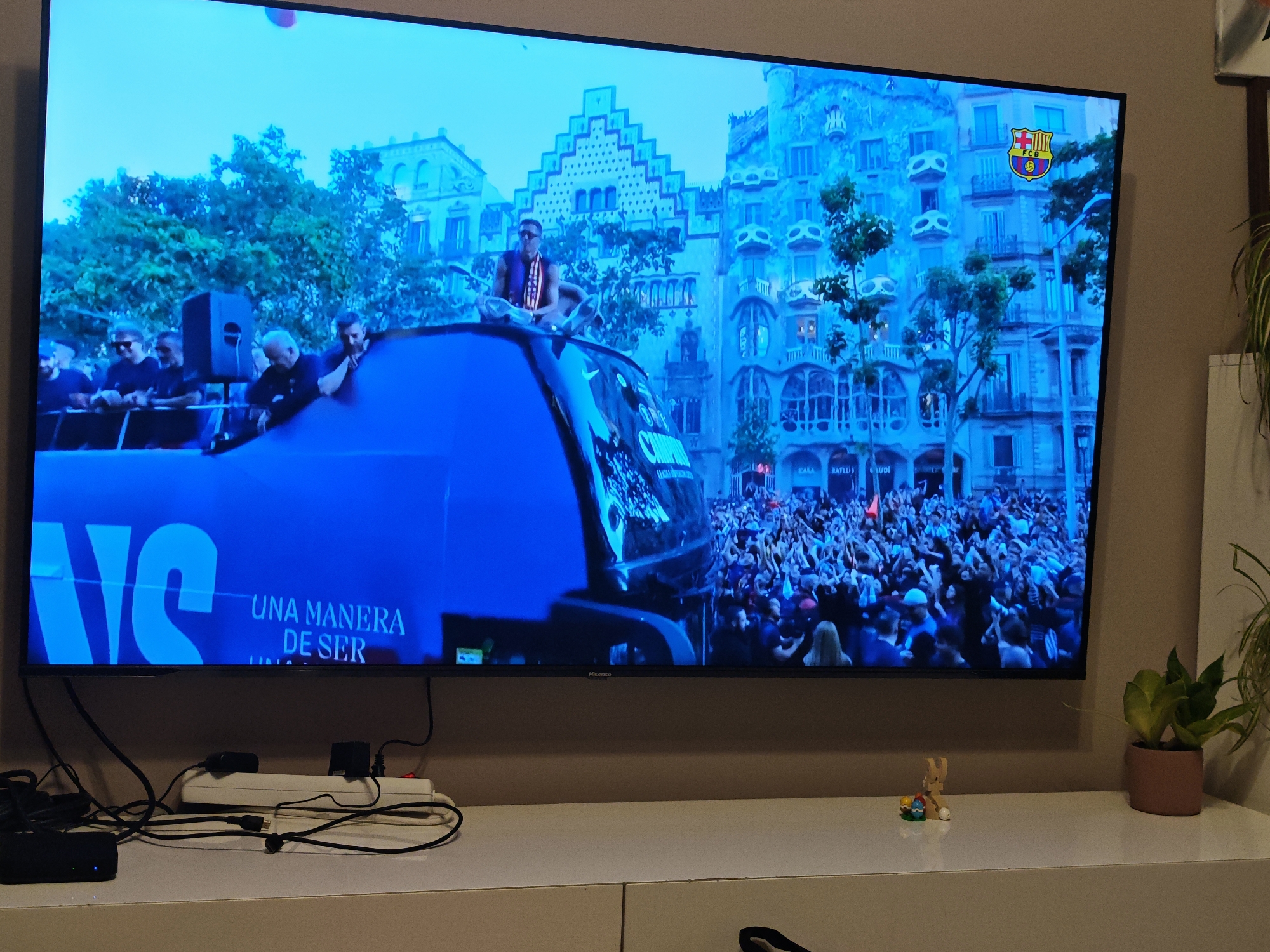 The image shows a television displaying a scene of a public celebration, likely a parade or rally. A large crowd is visible, with people raising their arms. In the foreground is a blue vehicle with text that appears to be "UNA MANERA DE SER". On top of the vehicle, there are several individuals, including one wearing a striped vest or scarf. Buildings are visible in the background, displaying architectural details. A crest or logo, potentially a sports team emblem, is displayed at the upper right of the screen. The TV is placed on a white console with a power strip and electronic devices connected. A small wooden figure and a potted plant are also present on the console. The scene displayed on the TV does not appear to be in Sofia, Bulgaria.