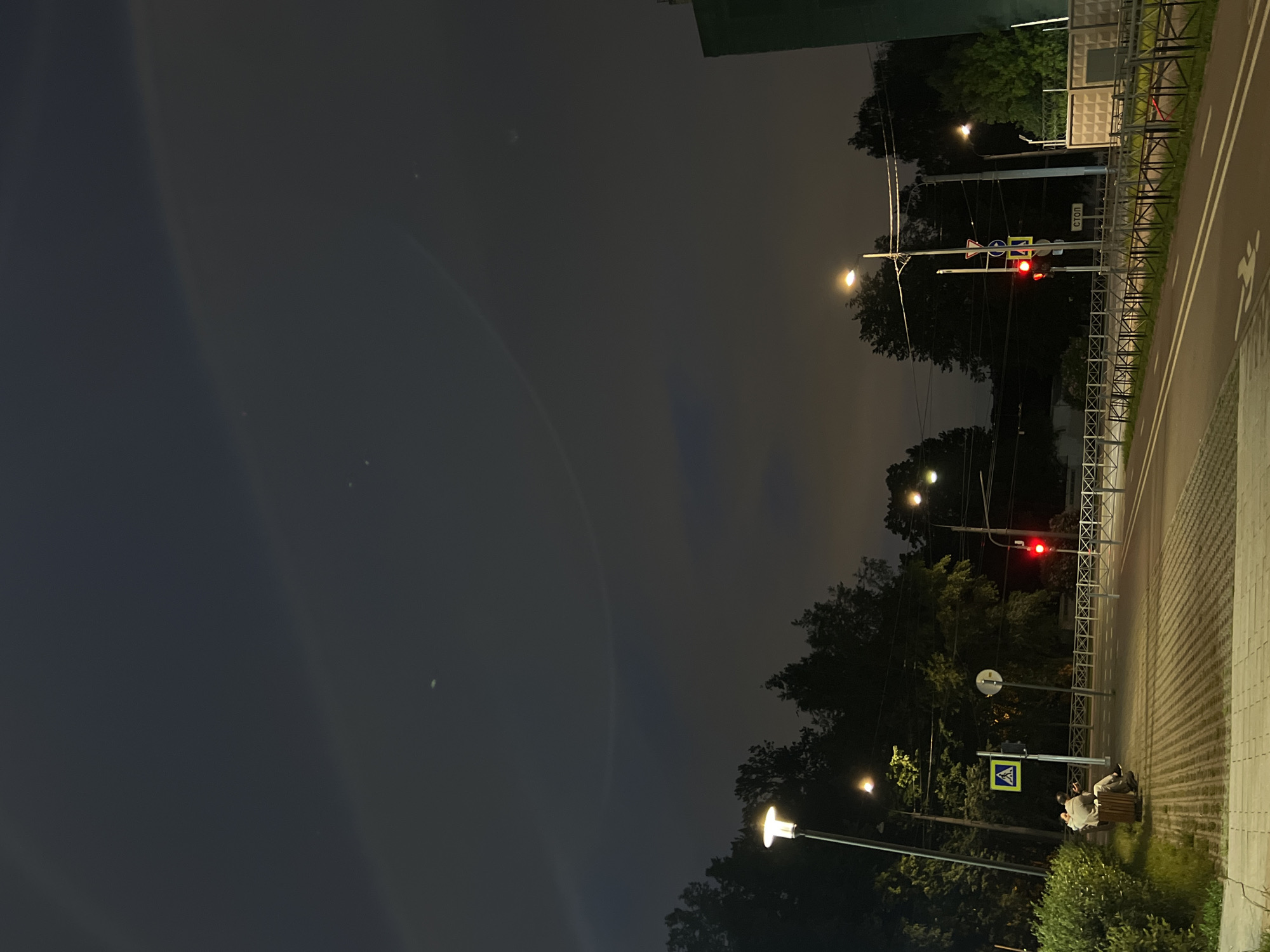 This image captures a nighttime scene on a street in Saint Petersburg, Russia, possibly near a residential area, judging by the presence of trees and a building. The sky is dark and cloudy, with a faint, arc-like light phenomenon visible, suggesting a halo around the moon or a similar atmospheric effect.
In the foreground, a pedestrian walkway is visible, made of light-colored rectangular paving stones. To the left, a person is seated on a wooden bench, facing away from the camera, looking towards the street. They are wearing dark clothing. Adjacent to the bench is a streetlight casting a bright, warm light downwards. A green bush is situated to the far left of the frame.
The street itself is paved with asphalt and has white lane markings. There are traffic lights visible; one on the right side of the street shows a red light, and another further down also appears to be red. Several traffic signs are also present, including a pedestrian crossing sign and a stop sign with the Cyrillic word "СТОП".
A metal fence with a diamond pattern separates the sidewalk from the street in some areas. Trees line the background, obscuring much of the view beyond the street. A section of a building, painted green, is visible on the far right of the image. The overall atmosphere is calm and quiet, characteristic of a city street at night.