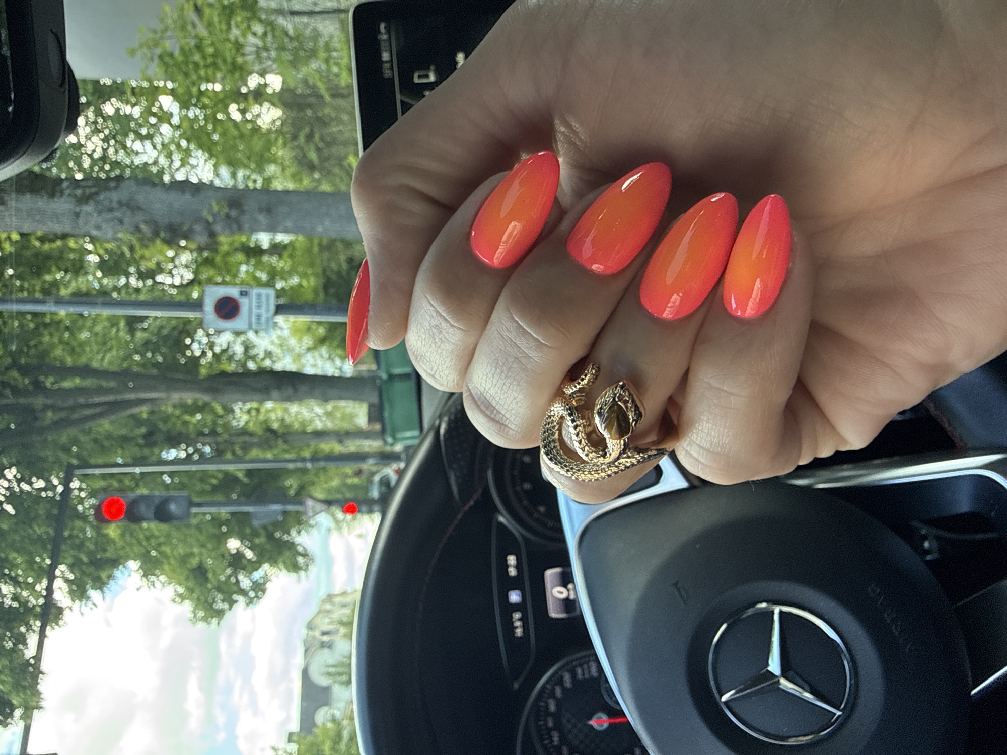 The image shows a hand with orange ombre nail polish and a gold snake ring, positioned over the steering wheel of a Mercedes-Benz. The background is blurry but appears to be a street scene with trees and traffic lights showing red. The scene is located in the city of Fontenay-aux-Roses, France. A traffic sign with a "no parking" symbol is visible behind the hand.
