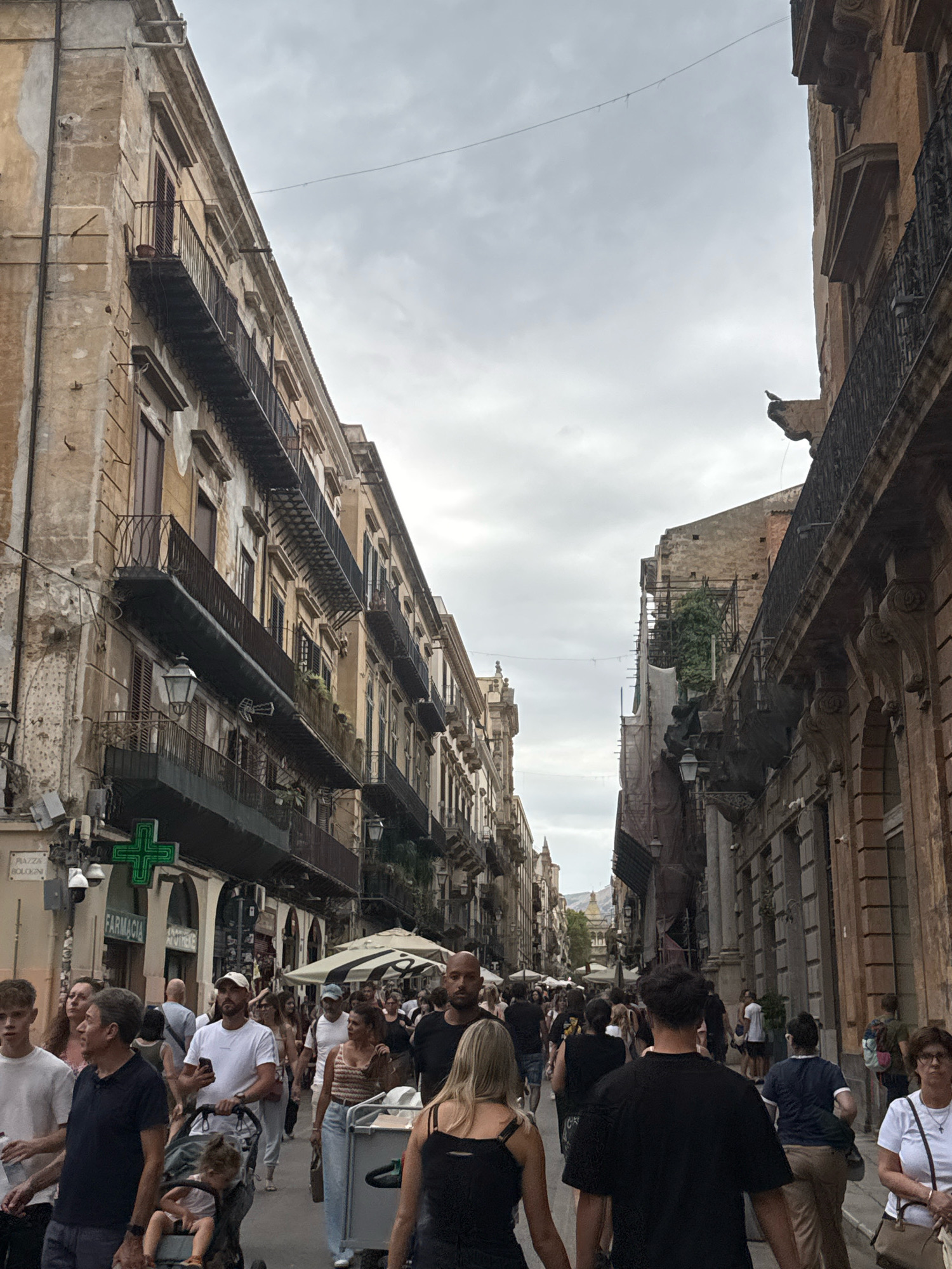 The image depicts a crowded street scene in Palermo, Italy, likely on Via Maqueda, a pedestrianized shopping street. The architecture features old, weathered buildings with balconies. The sky is overcast. A diverse crowd of people, including adults and children, are walking in both directions along the street. Some are pushing strollers. There are shops lining the street, with signs indicating businesses such as "FARMACIA" (pharmacy) visible. Umbrellas from outdoor seating areas are also present. The overall impression is a busy urban environment.