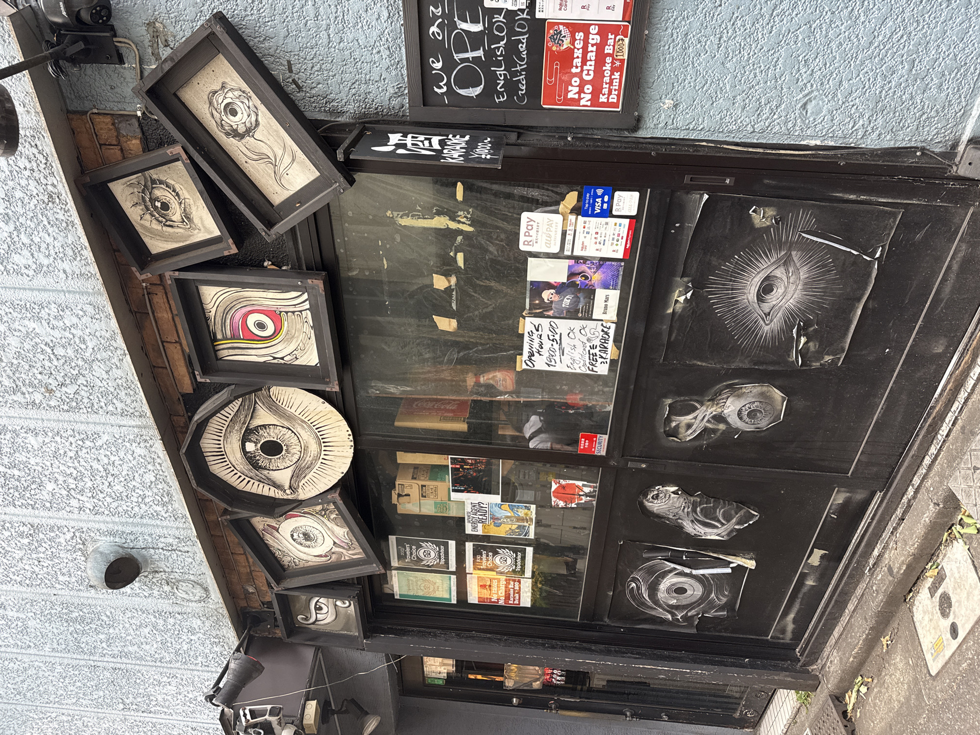The image shows the exterior of a bar in Shinjuku, Japan, called "Karaoke Bar". The bar features a dark storefront with large glass windows that display various posters and artwork, predominantly featuring stylized eye motifs. Above the windows, several framed artworks, also depicting eyes, are mounted on the building's facade. A black chalkboard sign on the right side of the entrance reads "we are OPEN" in white chalk, followed by "English OK" and "Credit Card OK". Below this, a red and white sign indicates "No taxes No Charge" and advertises "Karaoke Bar Drink ¥1000~". The doors of the bar are black and adorned with more large, black and white eye-themed decals. The surrounding building has a light blue, textured concrete wall. A metal pipe and vent are visible on the wall above the entrance, along with a security camera. The ground in front of the bar is paved, with a curb and some scattered fallen leaves. The lighting suggests it is daytime, possibly overcast. No people are clearly visible within the bar.