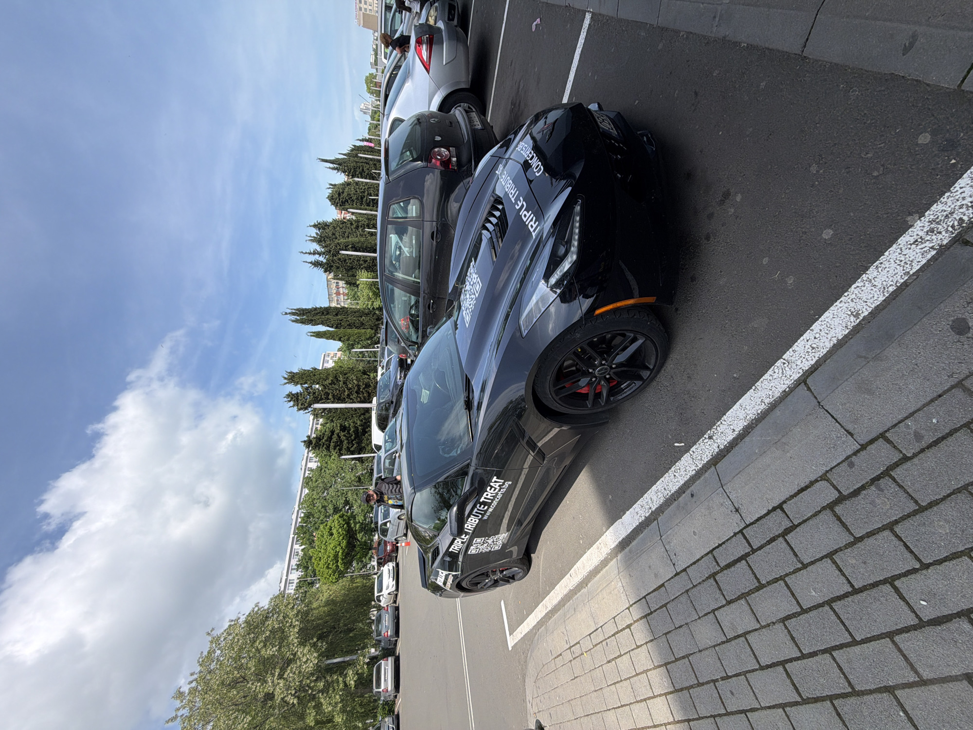 The image depicts an outdoor parking lot scene with a black Corvette parked near a curb. The car has red brake calipers and the words "TRIPLE TRIBUTE TREAT" written on its hood and side. Other vehicles, primarily compact cars in shades of grey and black, are parked nearby in parallel rows. There are people visible in the background near other parked cars. Trees and buildings are visible in the backdrop against a partly cloudy blue sky. The parking lot is paved, and the curb is made of gray paving stones. The location appears to be in Burgas, Bulgaria, indicated by the web address, of the car's design, as well as a building in the background that resembles architecture in Burgas.