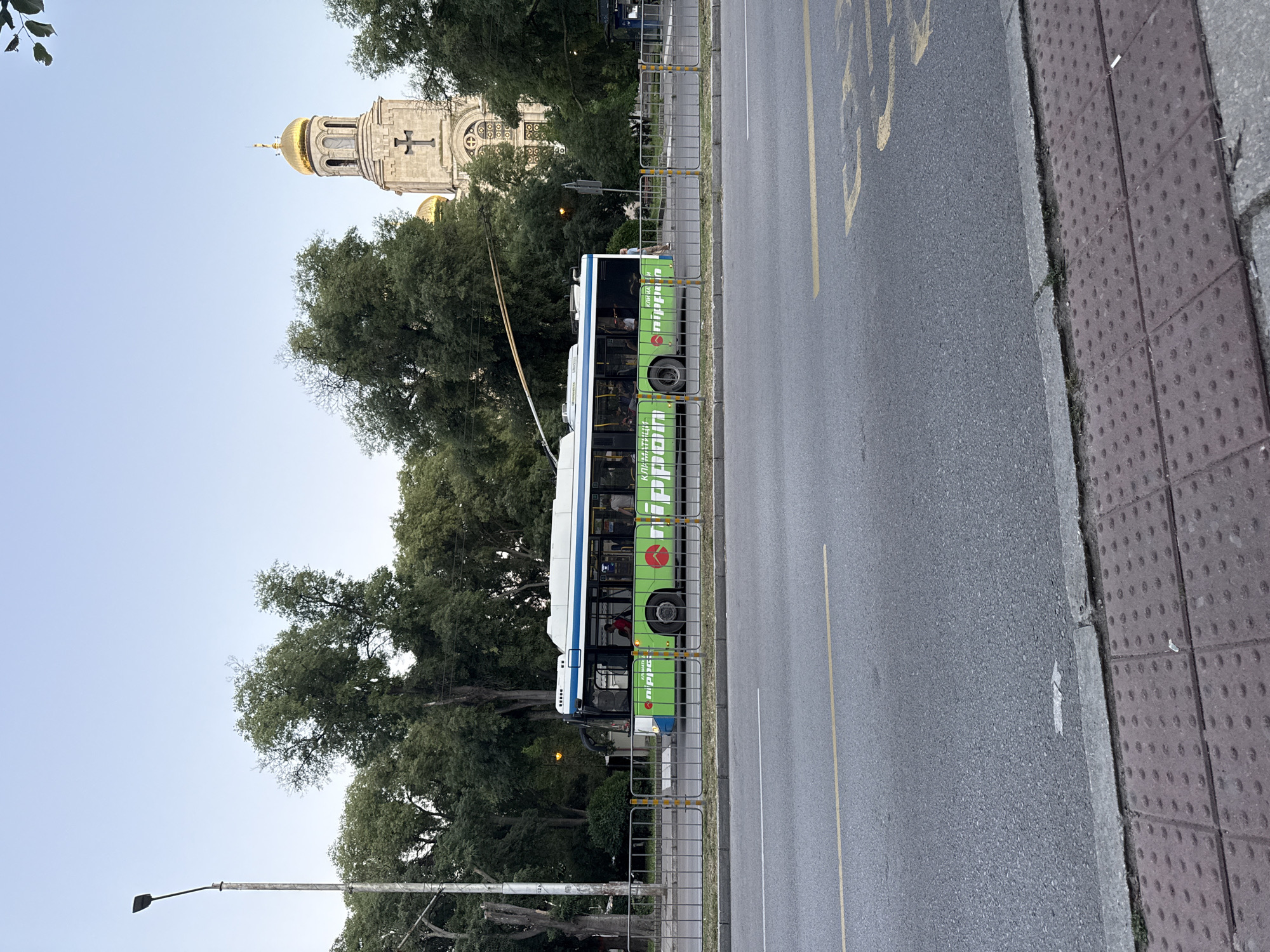 The image shows a street scene with a trolleybus and a building in the background. The trolleybus is green and white with text and logos on its side. Several people are visible inside the trolleybus. The building in the background appears to be a church with a golden dome and a cross. Trees line the street, and there is a sidewalk with textured paving stones. A street light is also visible. The image is taken from a low angle and appears to be taken during the day, likely late afternoon or early evening due to the lighting. The scene suggests an urban environment. The text on the bus is in Cyrillic script. One visible logo is a red heart shape.