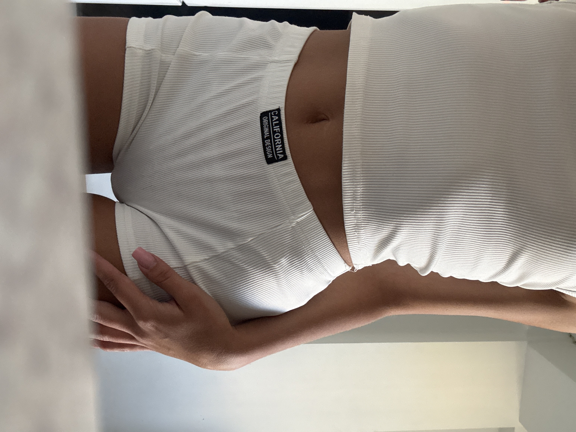 A person is shown from the waist down, wearing a white ribbed tank top and matching white ribbed shorts. The shorts have a black label with white text that reads "CALIFORNIA ORIGINAL DESIGN." The person's hand is resting on their hip. The background is out of focus but appears to be an indoor setting with a wall and a doorway. There is no information in the image to suggest this location is in Chișinău, Moldova.