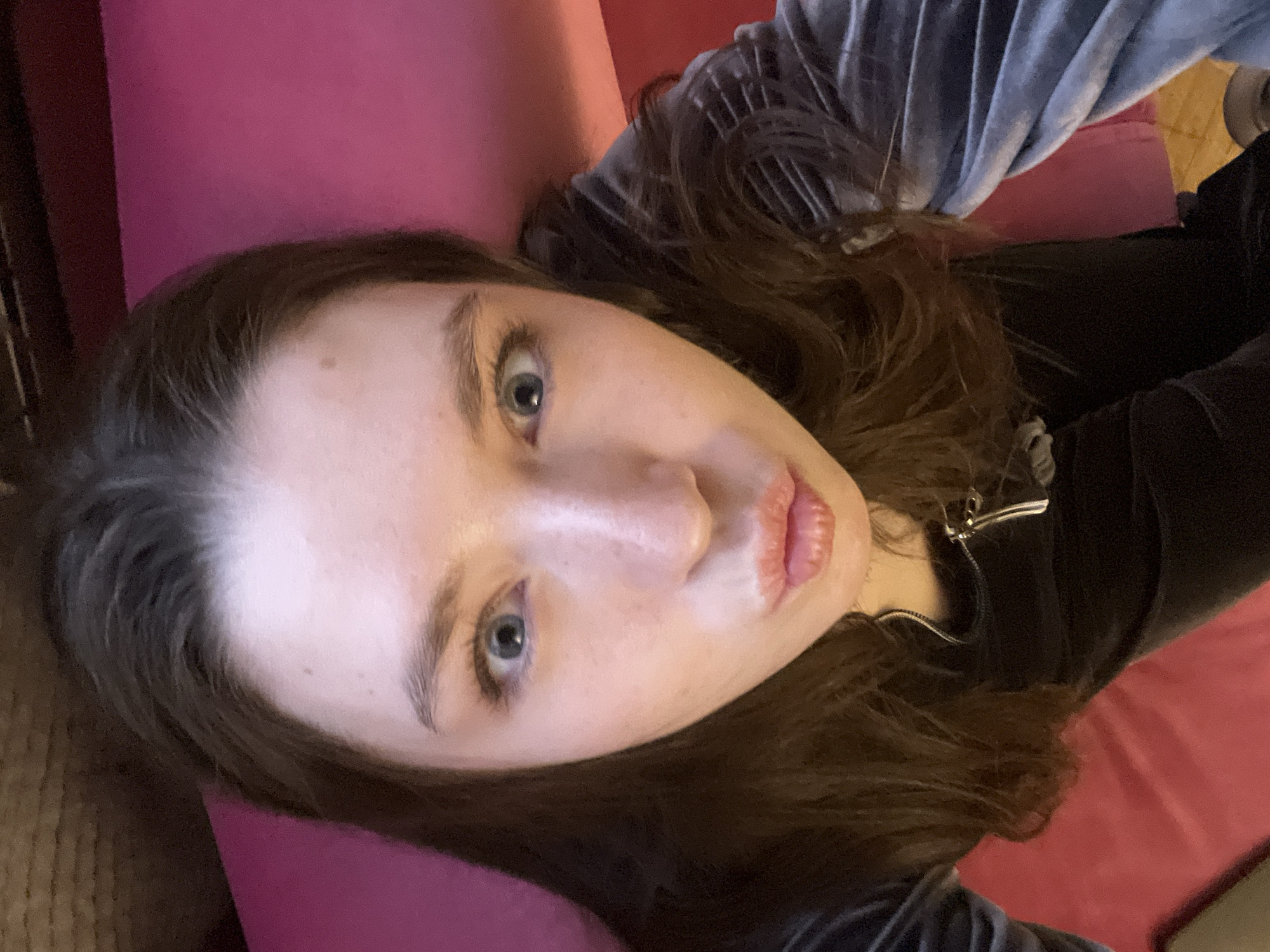 A woman with light skin, brown hair, and blue eyes is lying down on a red surface, possibly a couch or bed. She is wearing a dark-colored top with a silver zipper. A gray or blue velvet-like garment is visible on her upper torso. The background includes a dark-colored wooden structure.
The location is not discernible.