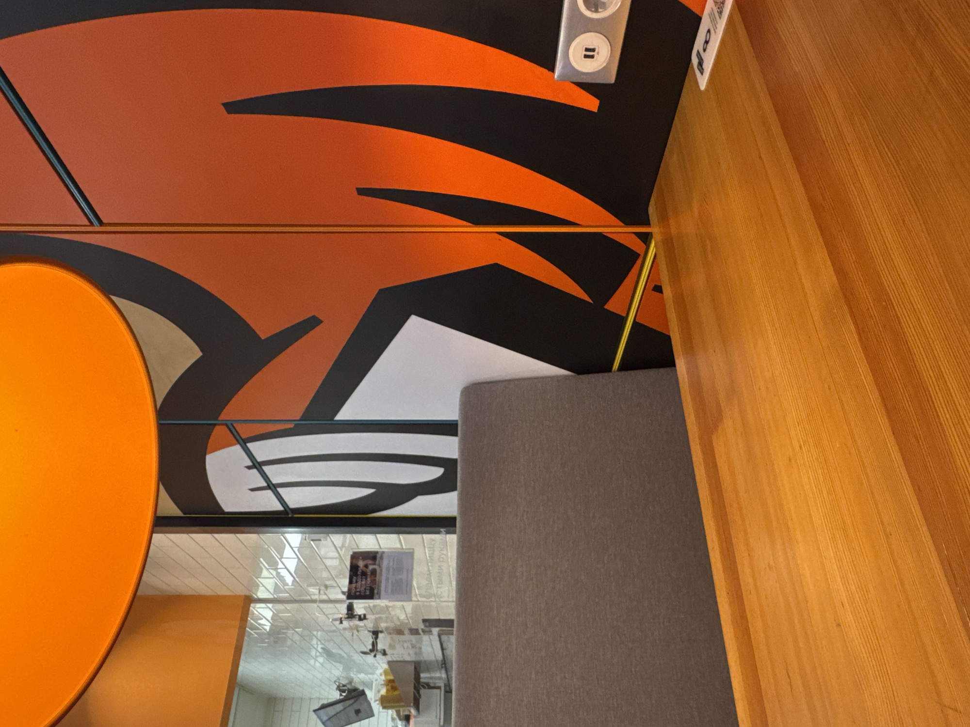 The image shows a partial view of an interior in what appears to be a commercial setting in Rostov-on-Don, Russia. A portion of a vibrant orange wall with stylized black graphics is visible, with a dual electrical outlet and a small paper label attached near the top right corner. Below, a light-colored wooden cabinet or wall panel runs vertically along the right side of the frame. To the left, an orange circular element, likely part of decor or signage, is cut off by the frame. A gray fabric-covered booth or seat back is positioned between the wooden panel and the background. In the background, a bright, tiled area is visible through a transparent barrier, possibly glass or plastic, with a glimpse of what looks like a kitchen or food preparation area. A small notice or advertisement with printed text is affixed to the transparent barrier. Overall, the scene suggests a modern or stylized dining area, potentially a restaurant or cafe.