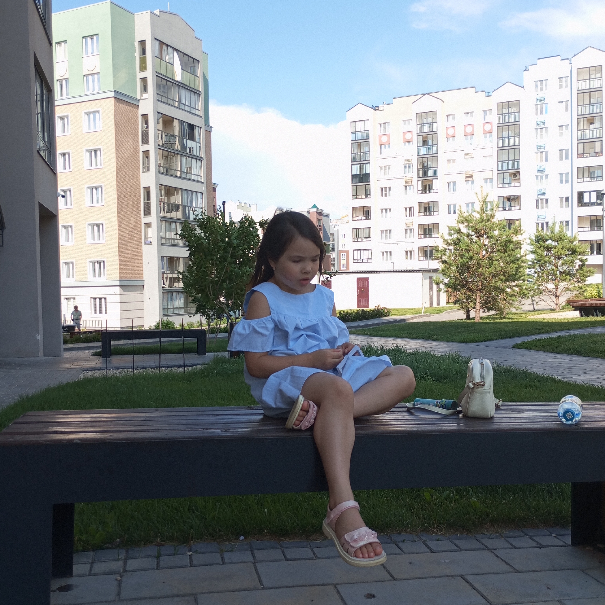 In Pervomaysky, Russia, a young girl is sitting cross-legged on a dark wooden bench in a courtyard. She is wearing a light blue off-the-shoulder dress and pink sandals. Her hair is dark and pulled back. In front of her on the bench are a white purse, a rolled-up tube or scroll, and a bottle of water. Behind the bench is a grassy area and a paved walkway. In the background, several modern apartment buildings of varying heights and colors are visible under a bright blue sky with scattered clouds. One building on the left has a light green upper section and a light yellow and brown lower section. To the right, there are taller, predominantly white buildings with multiple balconies. Two evergreen trees are planted in the courtyard.