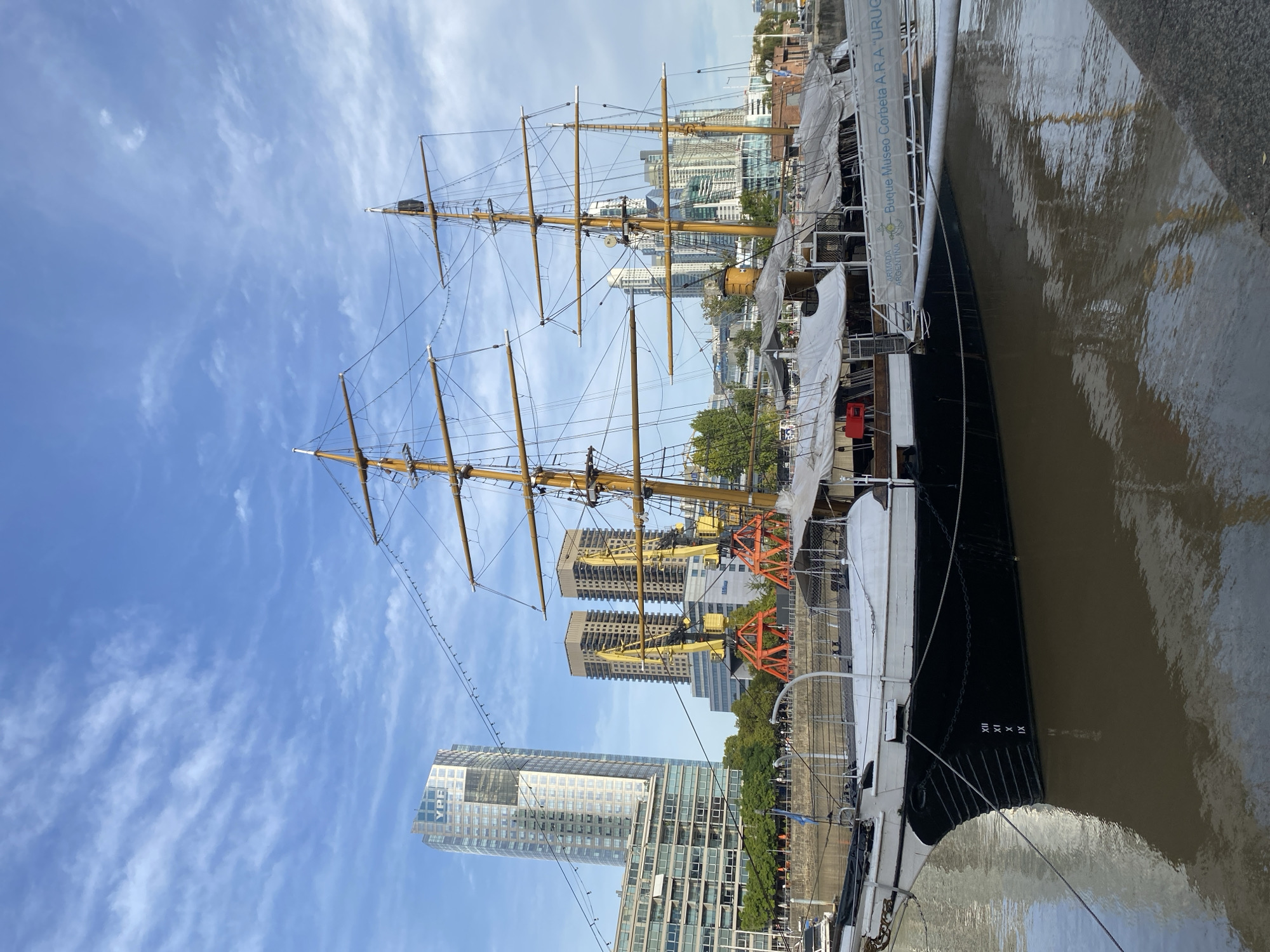 The image depicts a large, black and white tall ship docked in the city of Buenos Aires, Argentina, with the masts extending high into the blue sky. The ship's hull is reflected in the murky water, and the names 'Armada Argentina' and 'Buque Museo Corbeta A.R.A "Uruguay"' are visible along the side. Behind the ship, modern high-rise buildings dominate the cityscape. The YPF building is clearly visible among the other modern buildings. Along the dock, are cranes and a few trees and street furniture. The day is clear with some clouds in the sky. No people are prominently visible in the scene.