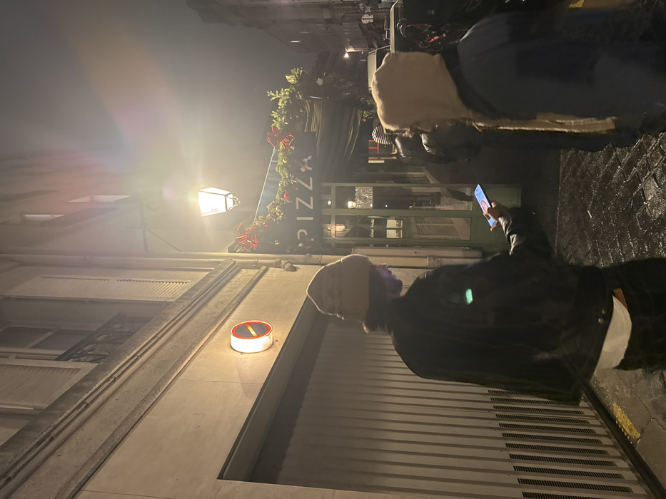 A nighttime scene reveals a streetscape with a "Pizza" sign adorned with greenery and red bows. A person in a dark jacket and beanie holds a phone, appearing to take a picture. A street lamp casts a warm glow, and a circular sign with a red center is visible on a building. The street appears wet and reflections shimmer on the surface. A second person with blonde hair is partially seen in the upper right.