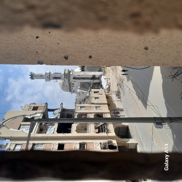 The image depicts a street scene in Khan Yunis, Palestinian Territory, characterized by extensive structural damage. In the foreground on the left, a multi-story building, appearing to be residential or mixed-use, displays significant destruction with large sections of its facade missing, exposing interior rooms and balconies. Debris hangs from upper floors, and clothes are visible on some balconies. A utility pole stands vertically in the left-center, with wires extending across the scene.
On the right side of the foreground, a textured, light-colored wall, likely part of a building, is blurred and exhibits multiple small, circular impact marks. The street in the mid-ground is an asphalt road, appearing relatively clear, with shadows cast across its surface. Two individuals, dressed in dark clothing, are walking away from the viewer on the right side of the street.
In the background, a mosque with a distinct dome and a tall, multi-tiered minaret stands prominently, also showing signs of damage, particularly around its base and surrounding structures, where rubble is visible. Other smaller, severely damaged buildings and piles of debris flank the street, particularly to the right and near the mosque. The sky is partially cloudy.
