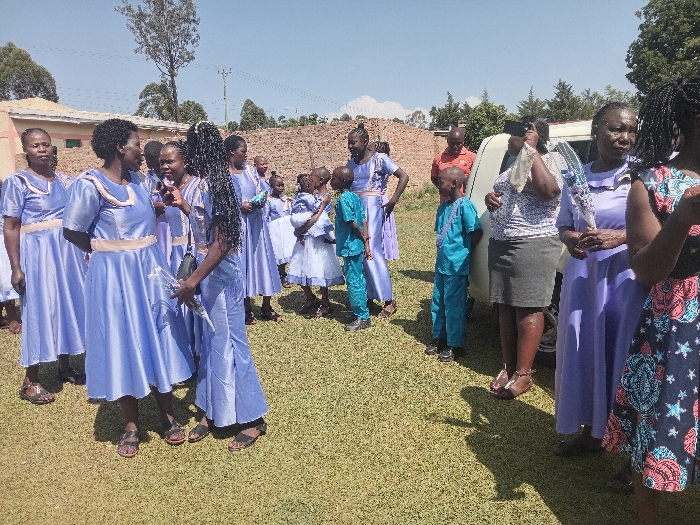 A group of people, predominantly women, dressed in blue and light-colored clothing, stand on a grassy lawn. Some are in matching dresses, while others are in various outfits. Children in similar blue outfits are also present. A white car is visible in the background, along with a few individuals, one in a red shirt. The scene appears to be an outdoor gathering or celebration.
