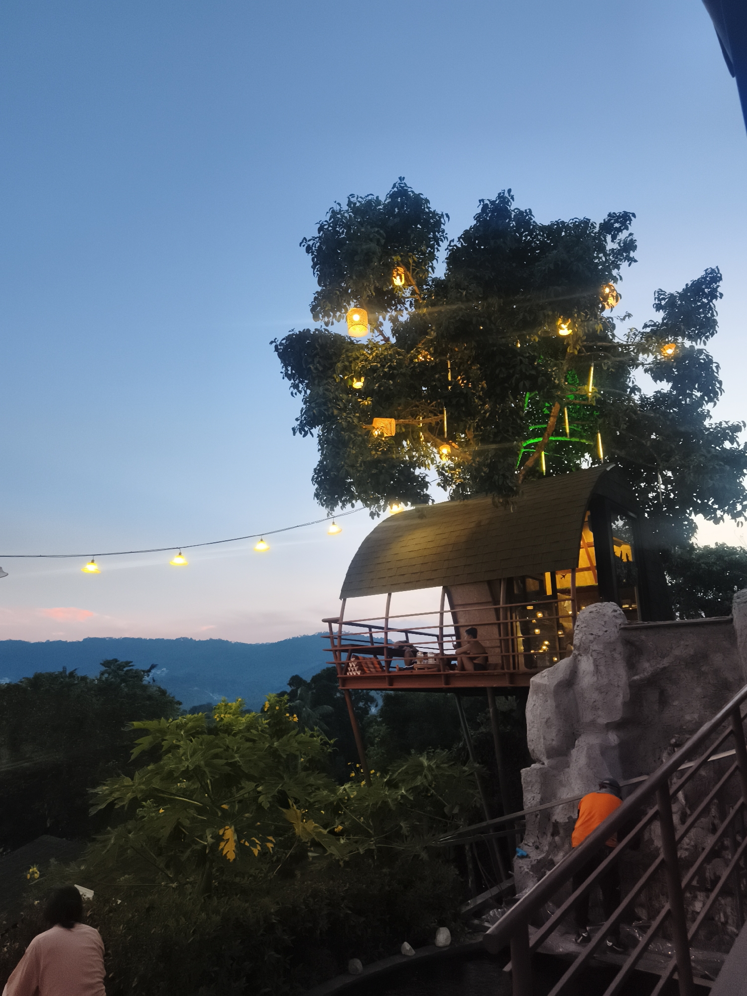 The image depicts an elevated treehouse restaurant in บ้านบางรักษ์, Thailand, set against a backdrop of distant mountains under a twilit sky. The restaurant is built around a large tree adorned with decorative lights, some of which are spherical and hanging from the branches. A string of lights stretches across the foreground, adding to the ambient lighting. People are visible on the deck of the treehouse, suggesting dining activity. In the foreground, a person wearing an orange shirt is ascending a set of stairs with metal railings adjacent to a grey faux rock structure. The surrounding vegetation is dense, indicating a natural or landscaped environment. A glimpse of another person is visible to the left.