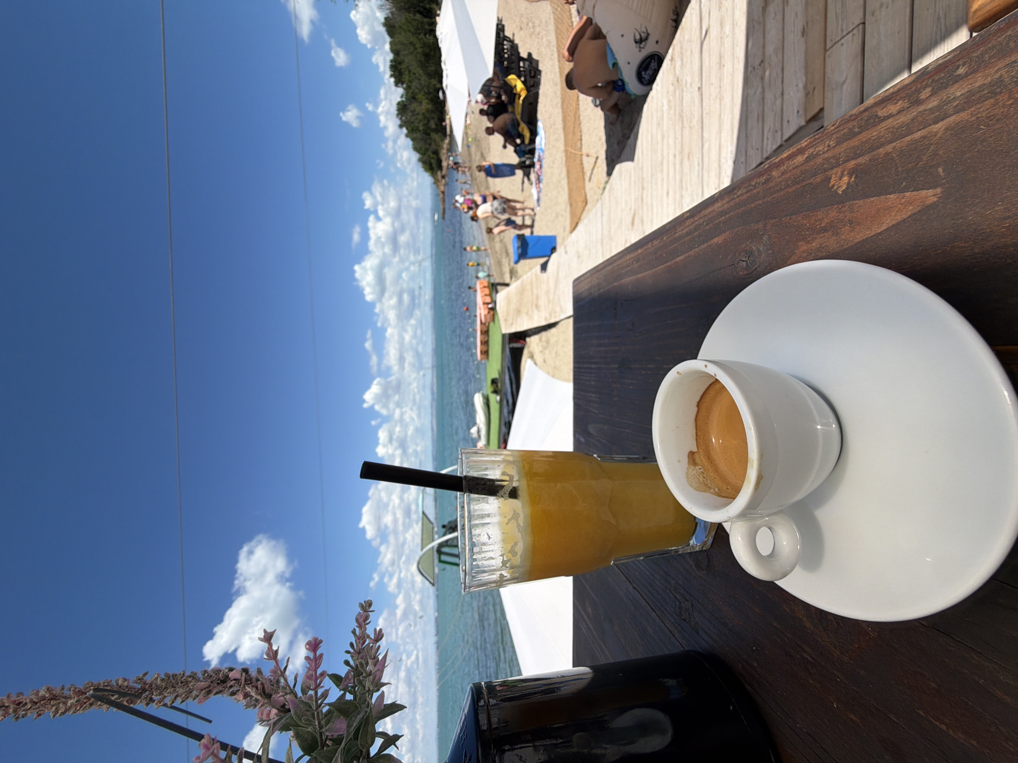 The image depicts a close-up of a wooden table in what appears to be a beachside cafe. On the table, two drinks are visible: a small white cup filled with espresso on a saucer to the right, and a tall glass of orange juice with a black straw to the left. In the background, a beach scene unfolds with people relaxing on lounge chairs under umbrellas. The sea is calm and blue, with a few boats and floating structures in the distance. The sky is clear blue with scattered white clouds. The location is identified as the beach in Burgas, Bulgaria, suggesting a summer day with recreational activities by the sea.