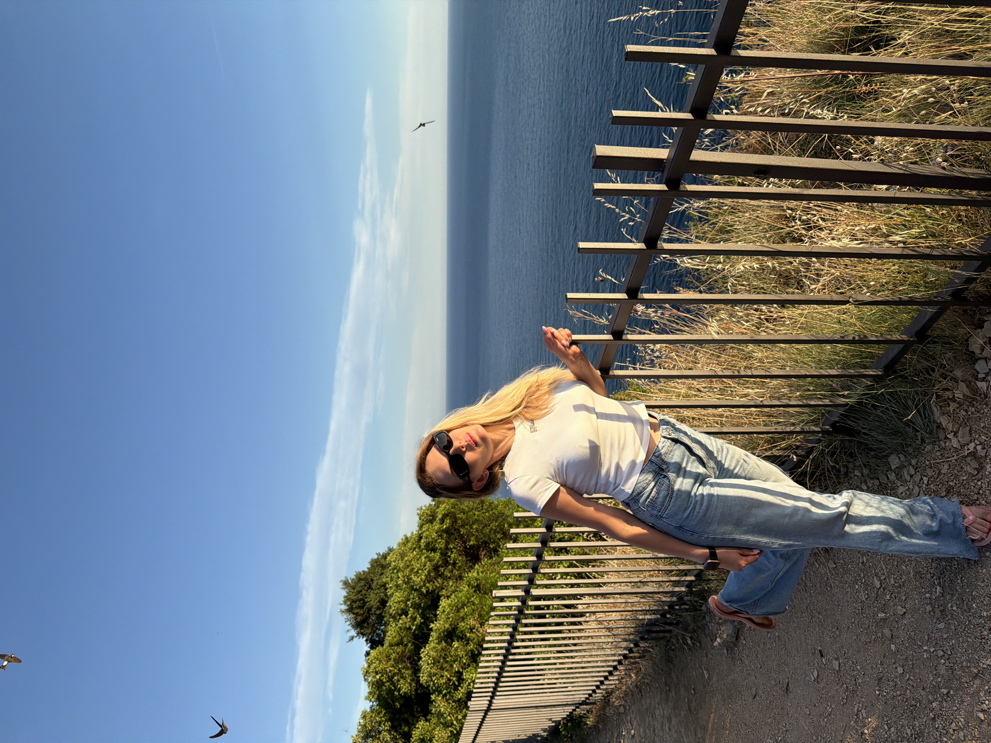 A blonde woman in a white t-shirt and blue jeans stands leaning against a black metal fence overlooking the Adriatic Sea. She is wearing sunglasses and flip-flops. The sun is setting, casting long shadows. The scene appears to be a scenic overlook in the city of Budva, Montenegro, likely near the historic Old Town, offering a view of the coastline and the vast blue sea under a clear sky with a few wispy clouds. Two birds are visible flying in the distance.