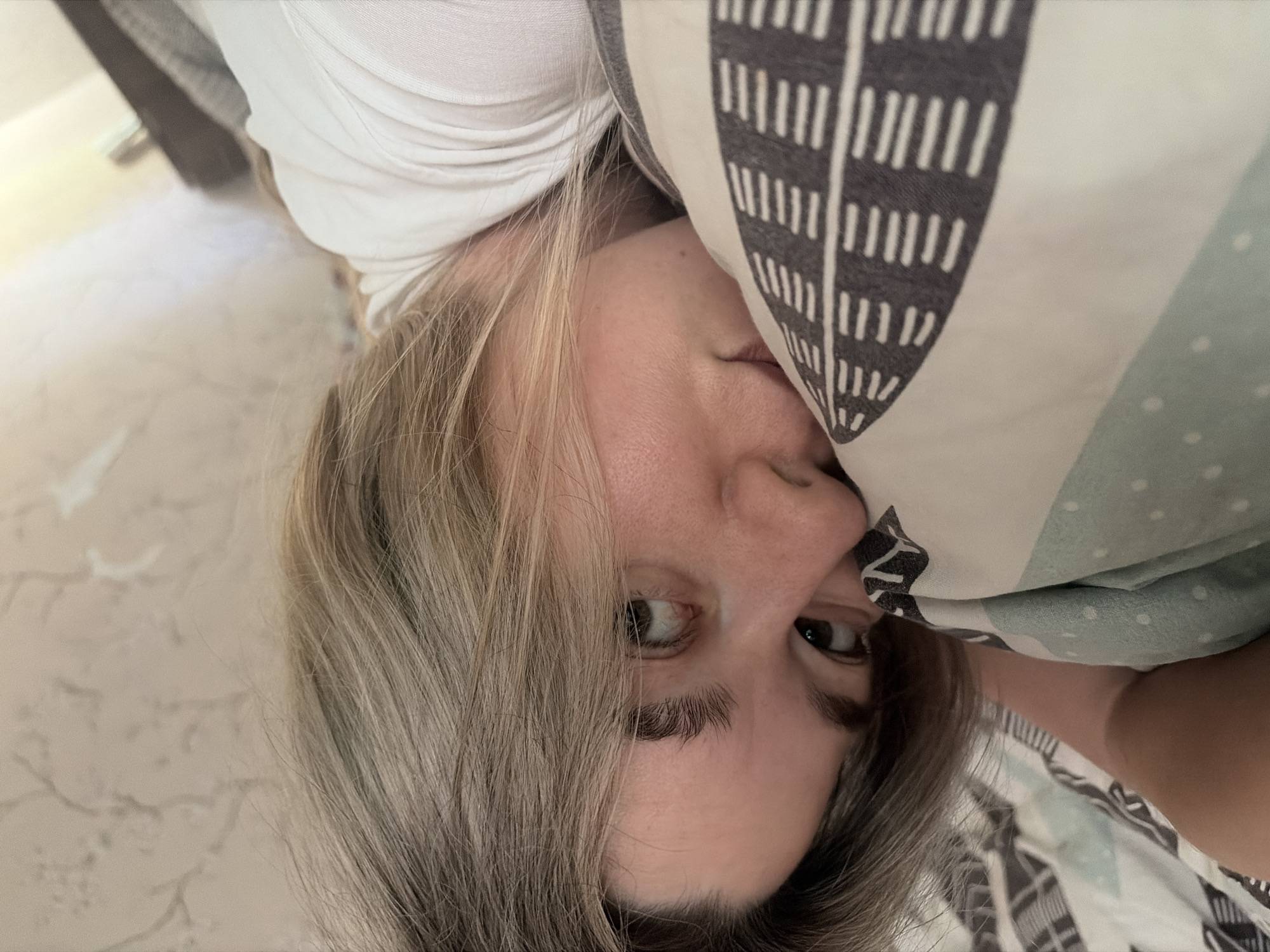 A woman is lying down, partially obscured by a patterned pillow with a light green section containing white polka dots and a white section with gray vertical lines. Her hair is a mix of blonde and gray. She is wearing a white shirt. The background suggests an interior setting, with a patterned wallpaper visible and a partially seen, dark-colored object behind her. The location is Chaikovsky, Russia.