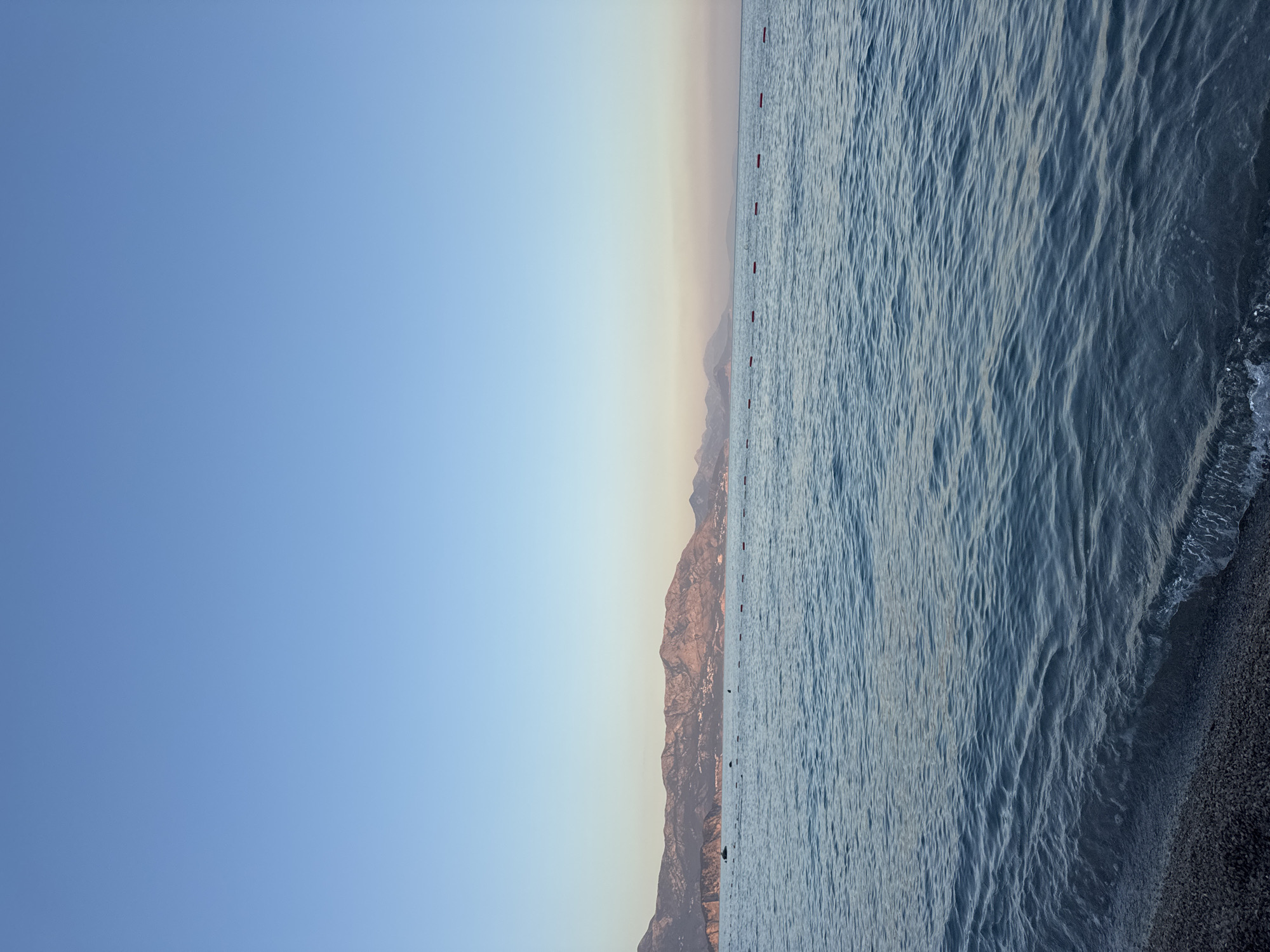 The image shows a wide expanse of the Adriatic Sea in Prijevor, Montenegro. The water is calm with gentle ripples. In the foreground, a pebbled shoreline is visible with small waves lapping at the edge. A line of red buoys stretches horizontally across the water, marking a boundary. In the distance, a range of mountains forms the backdrop, with a clear, pale blue sky above. The light suggests it is either early morning or late afternoon. No people or animals are visible in the scene. The overall impression is one of tranquility and natural beauty.