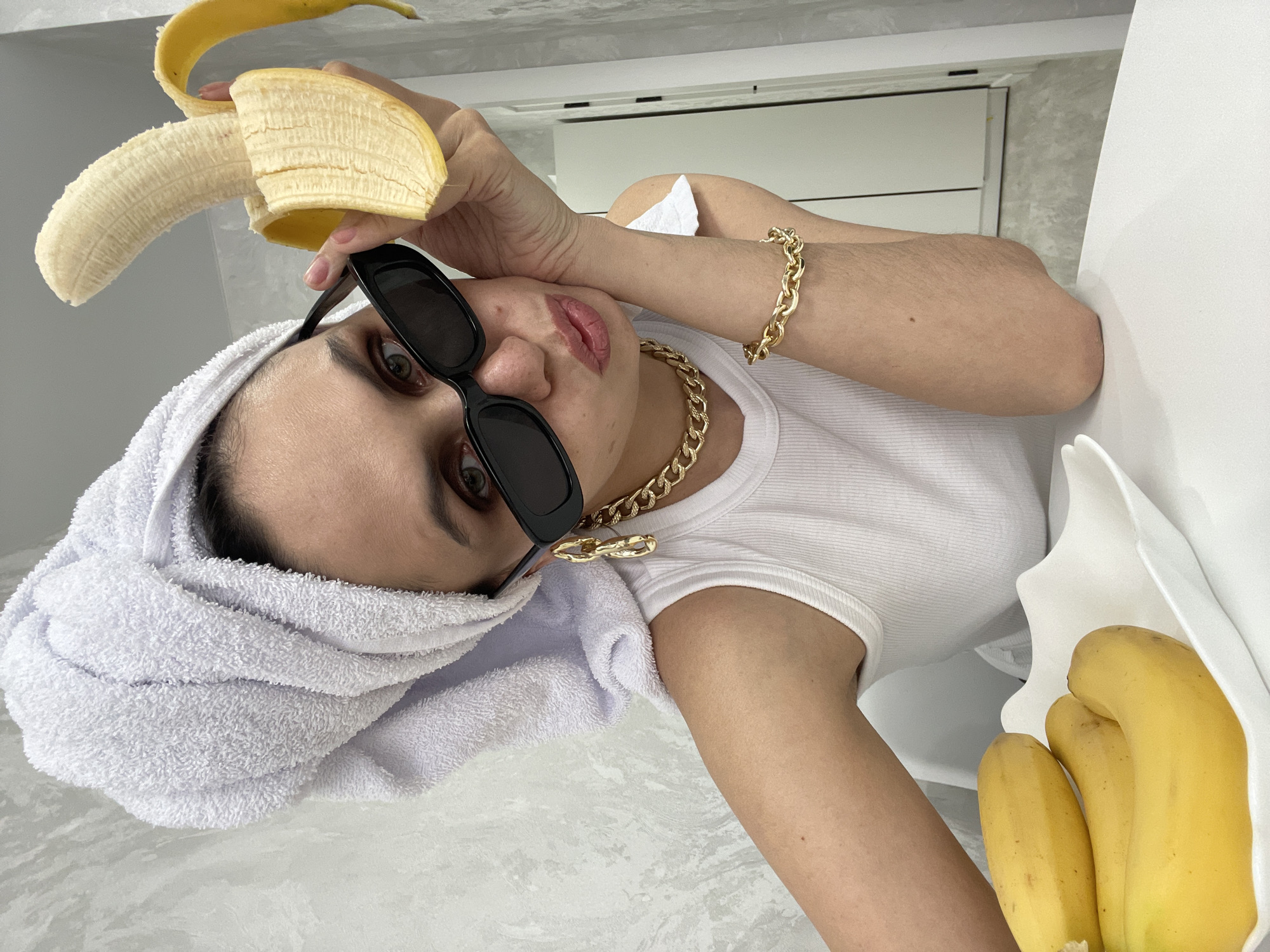 A woman wearing sunglasses and a towel on her head is holding a peeled banana. She is also wearing a white tank top and gold jewelry, and there are additional bananas in a white container next to her. The location is not discernable.