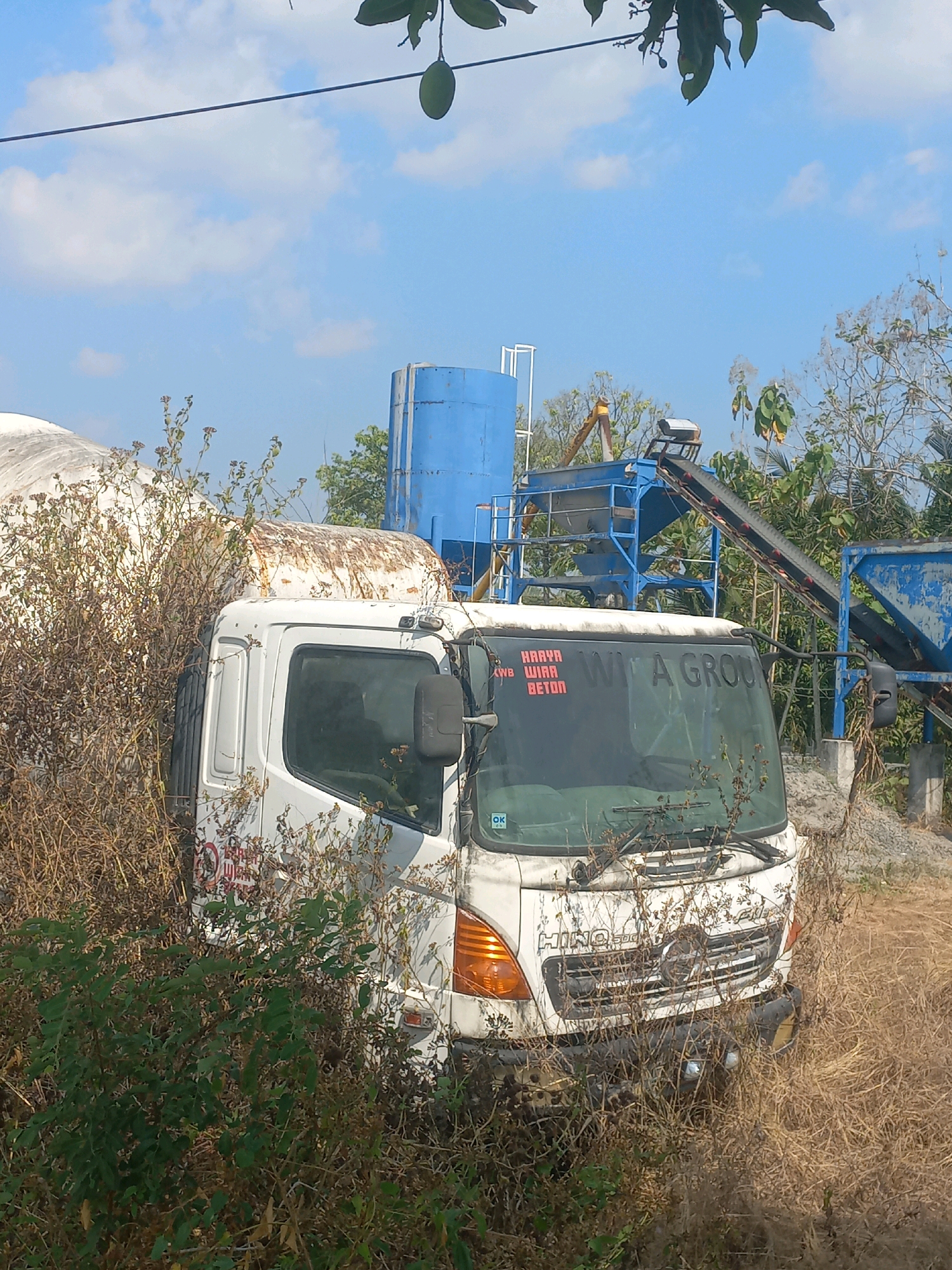 A white cement truck with red text on the windshield is parked amidst dry grass and foliage in what appears to be an industrial area. The truck's front is visible, with the brand name "HINO" clearly displayed on the grille. In the background, there are industrial structures, including a large blue silo and a conveyor belt system, suggesting a cement production or processing facility. The sky is blue with scattered clouds. The overall scene depicts an abandoned or disused industrial setting.