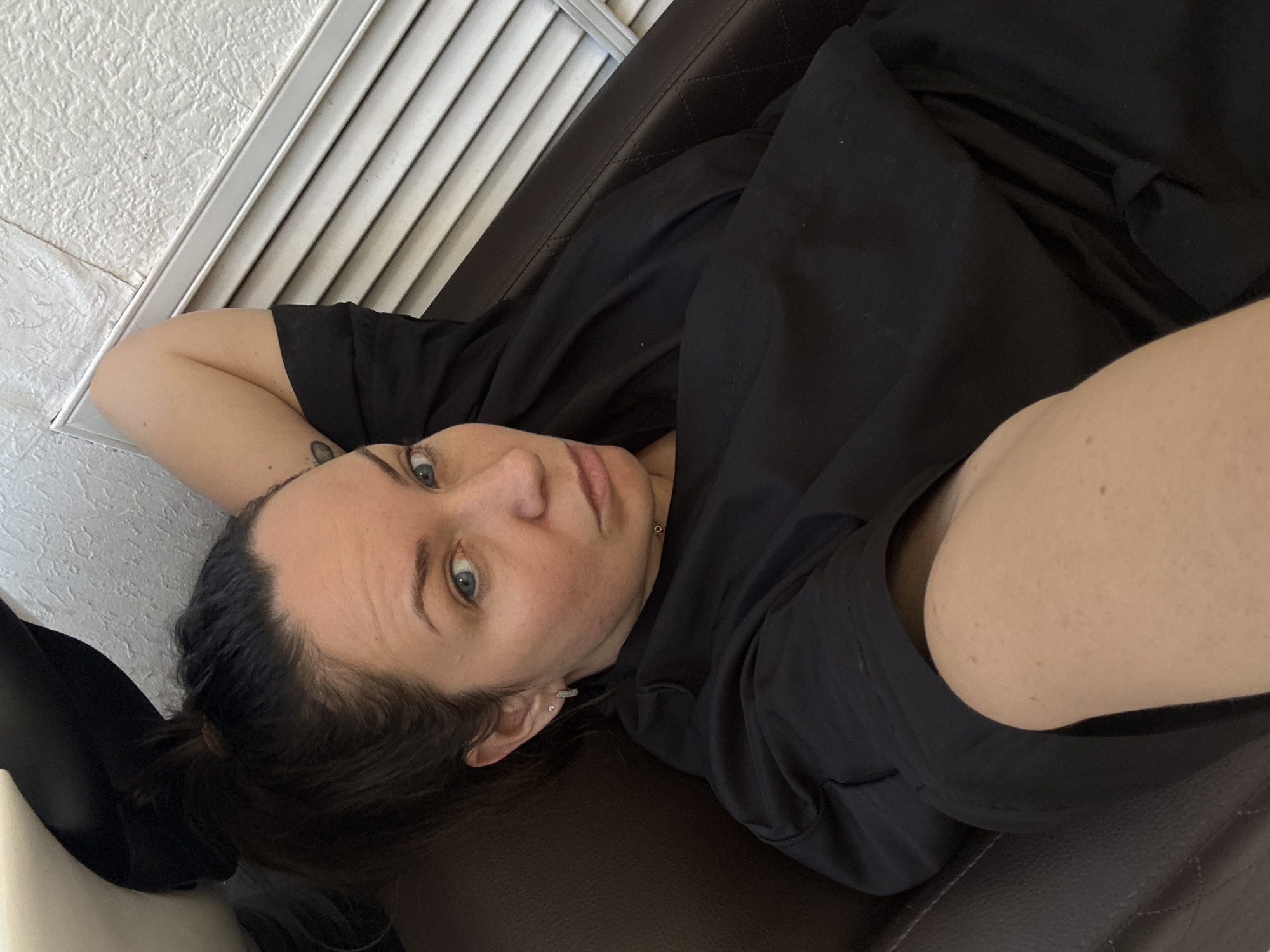A woman lies on a brown quilted sofa in a room with white textured walls and a white slatted window covering. She wears a black shirt and has dark hair pulled back into a bun. Her left arm is bent and raised behind her head. She looks directly at the camera with light-colored eyes. The location is unknown, and there is no evidence to suggest it is in the city of Chaikovsky, Russia.