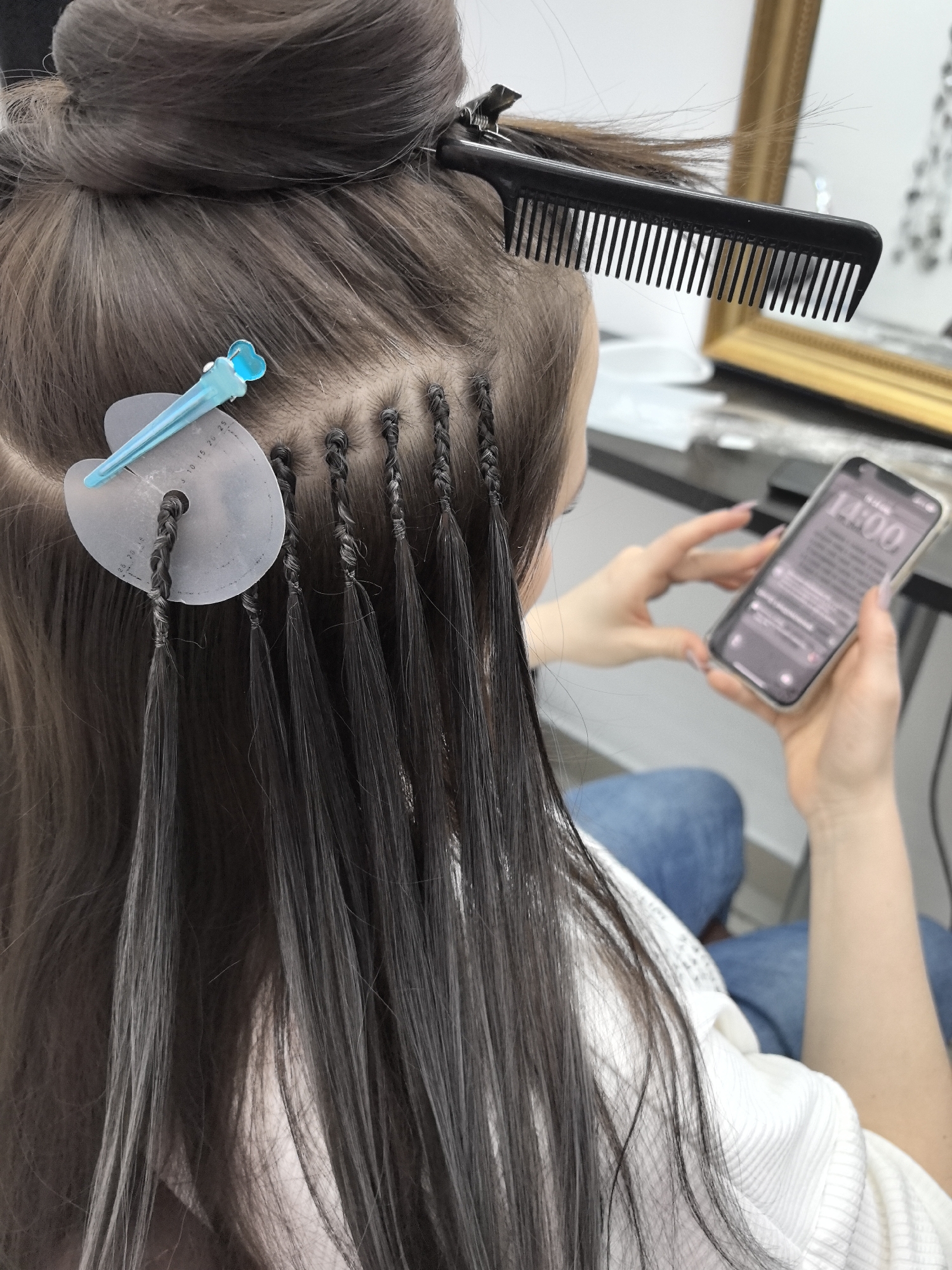 An overhead, slightly angled shot captures a woman undergoing hair extension application in what appears to be a salon in Chaikovsky, Russia. Her dark brown hair is partially tied up in a bun on the crown of her head, secured with clips and a black comb. Several individual hair extension strands are visible, meticulously braided into her natural hair close to the scalp, with the extended hair flowing downwards. One section is currently being processed, with a clear, circular hair protection shield positioned against her scalp, held by a light blue, heart-shaped clip. This shield features numbered markings, including '5, 10, 15, 20, 25'.
The woman is seated, wearing a white ribbed top and blue jeans, with her back mostly to the viewer. She is actively engaged with a white-cased smartphone, holding it in both hands and looking at its screen. The phone's display prominently shows '14:00'. The background is blurred, revealing a gold-framed mirror and a surface that resembles a salon station. The activity clearly depicts the process of hair extension application, with the client occupying herself during the procedure.