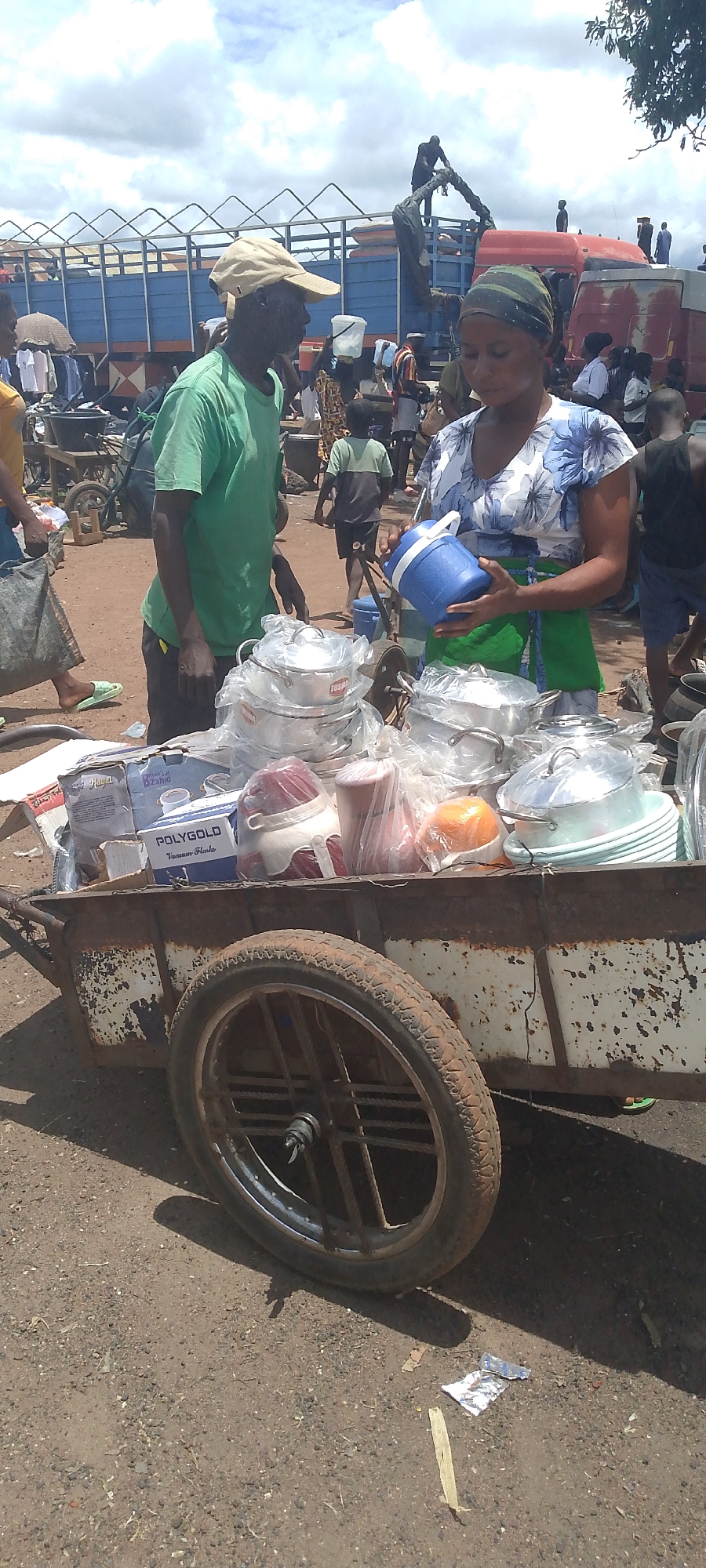 In Tor-Donga, Nigeria, a man wearing a tan cap and a green t-shirt stands next to a cart filled with kitchenware. A woman in a blue and white patterned dress and a headscarf holds a blue insulated container. The cart contains stacks of metal pots and pans, some wrapped in plastic, and boxes of "POLYGOLD Vacuum Flasks". The ground is a mixture of dirt and gravel, with debris scattered around. In the background, several trucks and people are visible, suggesting a busy marketplace or loading area. The sky is partly cloudy.