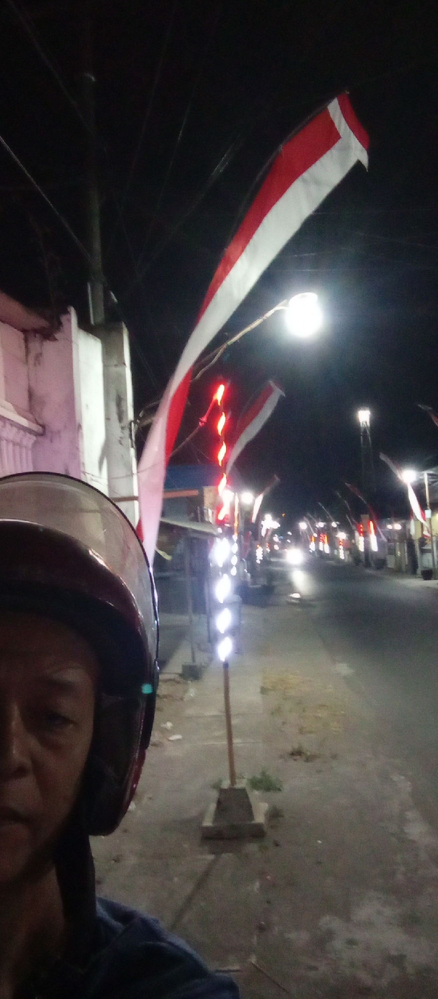 The image is a low-light, slightly blurry photograph taken at night on a street in Kediri City, Indonesia. The perspective is from a person wearing a red and black helmet, with their face partially visible in the foreground on the left.
The street appears to be paved and is illuminated by streetlights and decorative lights. Red and white flags, likely the Indonesian flag, are prominently displayed and appear to be strung across the street or along the sides. Some of these flags are caught by the wind and are waving. There are also vertical strings of bright white lights, possibly LEDs, along the side of the road. In the background, there are storefronts or buildings lining the street, and a vehicle's headlights are visible in the distance, suggesting traffic. The overall impression is of a festive or decorated street scene at night.