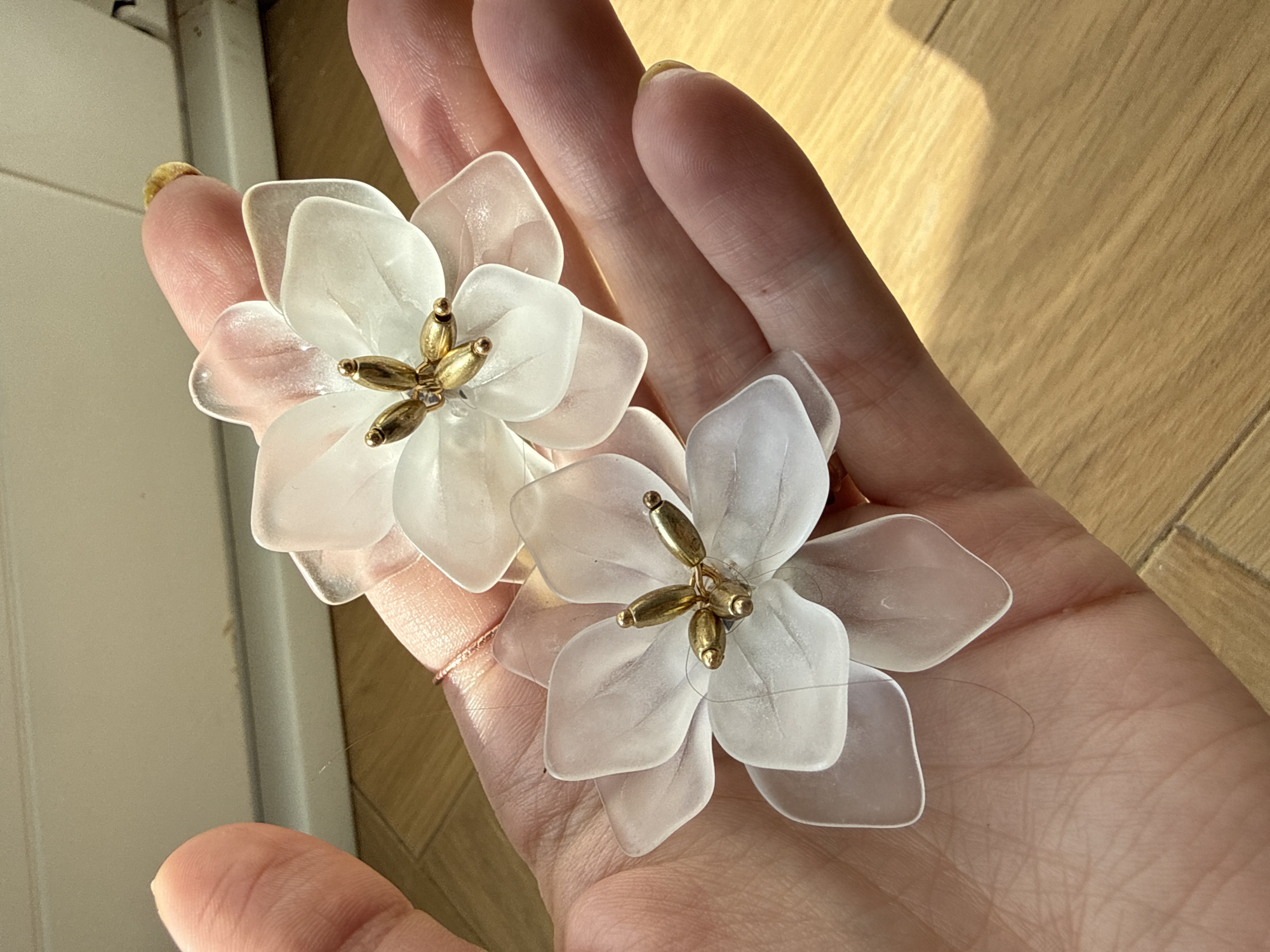 A person's hand holds two white, translucent flower-shaped earrings with gold-colored accents at the center. The background includes a light-colored wooden surface. The location is not discernible from the image.