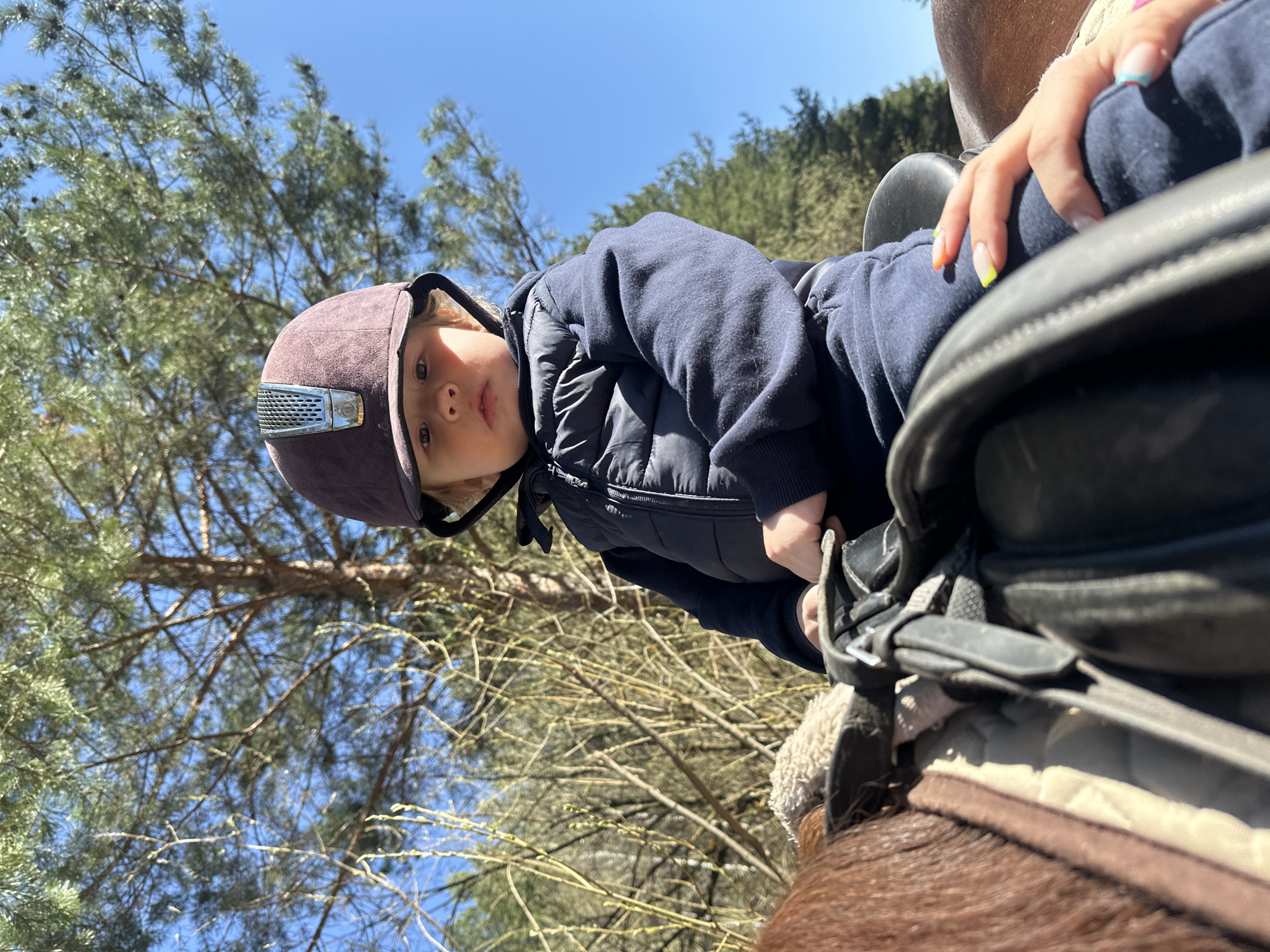 A young child wearing a burgundy riding helmet and navy blue outerwear is sitting in a saddle on a horse. The saddle is black leather and has a tan-colored pad. The child is looking directly at the camera. Another person, likely an adult, is partially visible and sitting behind the child, with a hand resting on the child's leg. The hand has painted nails, some with light-colored tips. The background includes trees and a blue sky. The location is outdoors, potentially near a wooded area. There is no information to support the image was taken in the city of Русский Вожой, Russia.