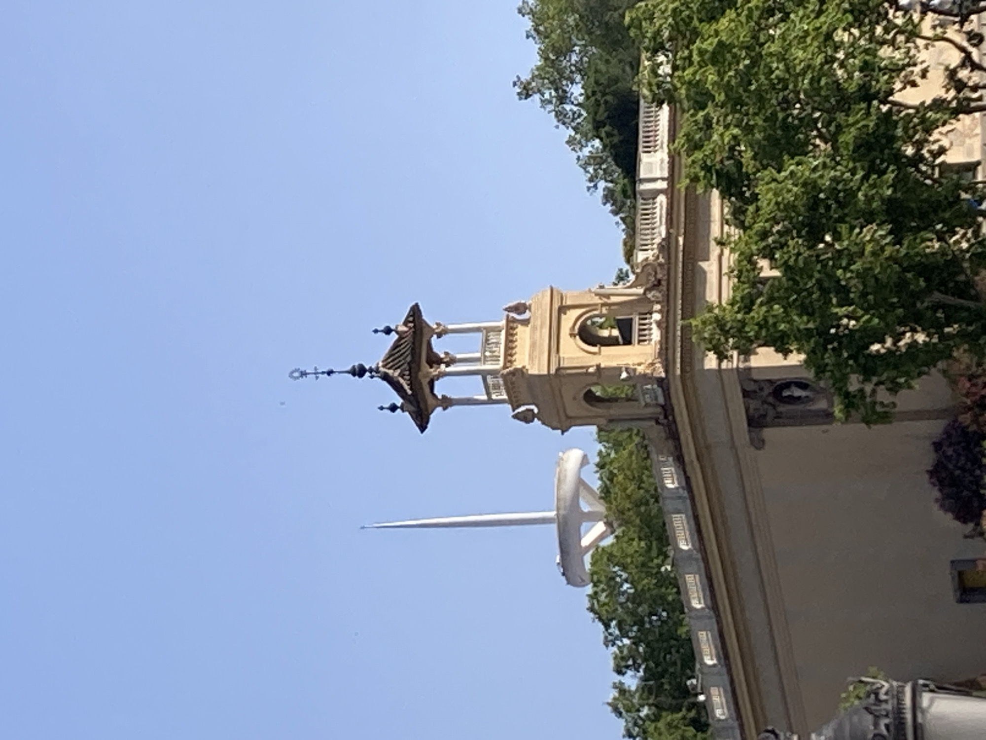 The image depicts a bright, clear day in Barcelona, Spain, with a view of the Montjuïc Communications Tower, also known as Torre de Comunicaciones de Montjuïc or Torre de Collserola, in the background. This distinctive white tower, a landmark of the city, features a tall, slender spire rising from a circular base supported by angled struts. In the foreground, the top portion of a light-colored building with classical architectural elements is visible. This building has a decorative balustrade along its roofline, and a prominent bell tower with a tiled roof and an open belfry is situated on its corner. The bell tower is adorned with ornate detailing and topped with a cross. Lush green trees surround the building, obscuring much of the lower structures and suggesting a park-like setting. The sky is a clear, vibrant blue, indicative of fair weather. No people are clearly visible in the frame, but the scene conveys a sense of historical architecture juxtaposed with modern infrastructure.