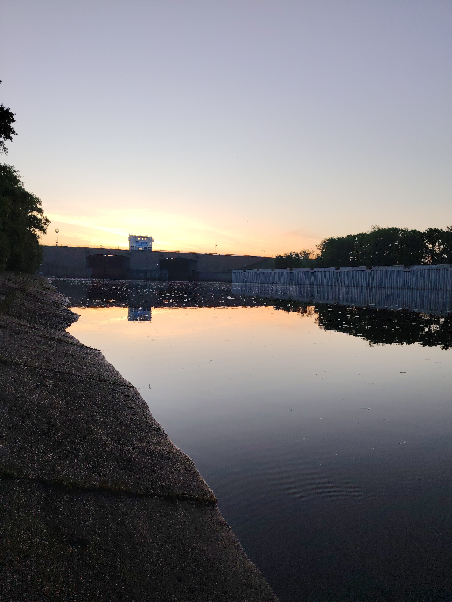 The image depicts the Kama River in Chaikovsky, Russia, at dusk. A concrete embankment forms the left foreground, leading to a calm expanse of water that reflects the sky and shoreline structures. In the background, a dam structure with a control tower is visible. A line of trees borders the dam on the right. The sky exhibits a gradient from pale yellow-orange near the horizon to a pale blue-gray above. Small ripples disturb the water's surface in the foreground.