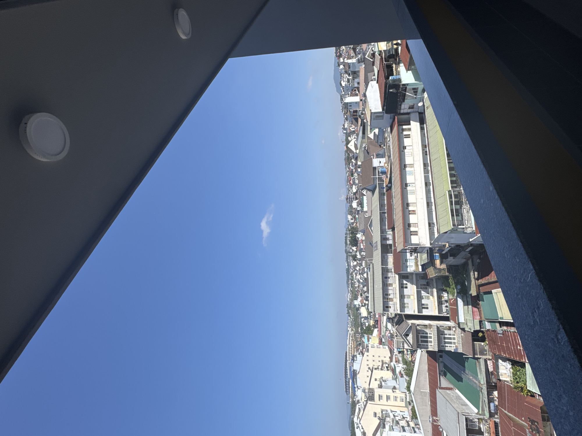 The image captures a vibrant urban landscape in Lâm Viên - Đà Lạt, Vietnam, viewed from an elevated perspective, possibly a balcony or a high window. The overall scene is dramatically tilted, presenting the city as a dense vertical strip against a vast blue sky.
The upper-left portion of the image is dominated by a clear, azure sky, indicating a bright and sunny day. A single, small, wispy white cloud floats near the center. This strong daylight suggests the photograph was taken during midday or early afternoon.
On the right, the cityscape stretches into the distance, revealing a tightly packed array of buildings. These structures appear to be mostly residential, with a diverse mix of architectural styles and roof materials, including corrugated metal roofs in shades of red, brown, green, and grey, as well as white and grey tiled roofs. The buildings vary in height, from smaller dwellings to multi-story apartments, hinting at the hilly terrain characteristic of Đà Lạt. The furthest visible structures blend into a hazy horizon, where faint outlines of mountains or distant hills can be perceived.
The view is framed by elements of the building the photographer is in. To the left, a dark grey ceiling or soffit is visible, featuring two modern circular flush-mount light fixtures. On the right, a textured, dark blue-grey vertical beam or wall serves as a partial border to the urban panorama. There are no people or discernible text visible in the image.