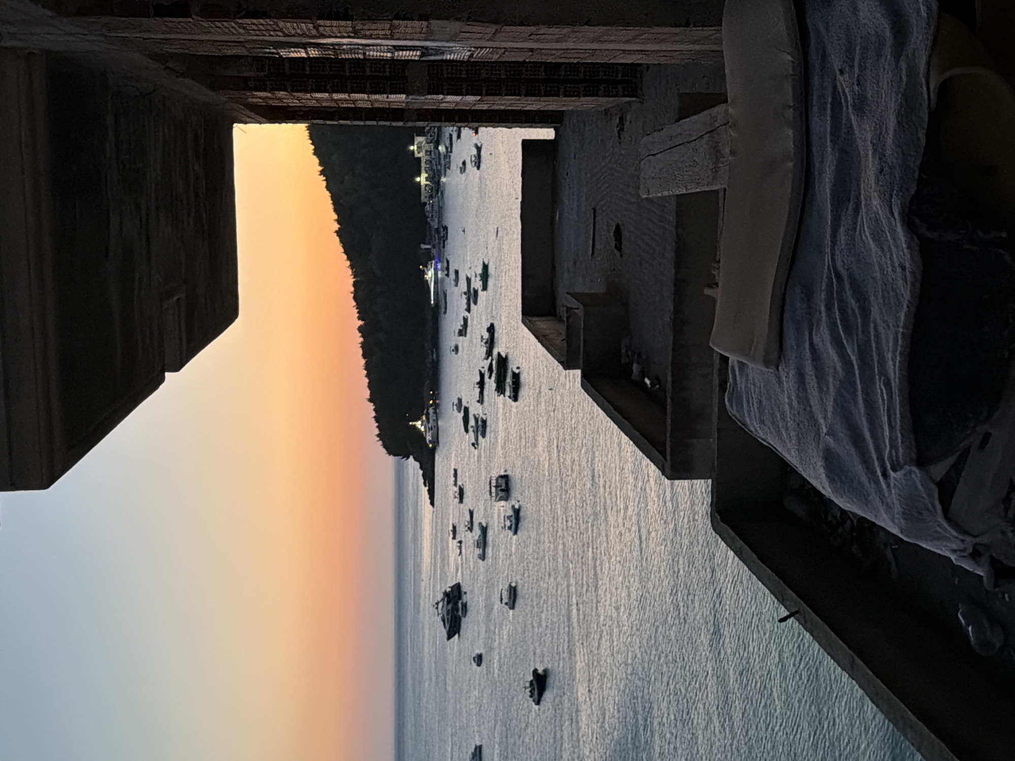 The image shows a view of the sea and coastline at sunset, with numerous boats anchored in the water. In the foreground, there is the edge of a partially constructed or abandoned building. The building's concrete structure is visible, with exposed rebar and unfinished surfaces. The sea is calm, reflecting the warm colors of the sunset sky, which transitions from pale blue at the top to a vibrant orange and pink near the horizon. A tree-covered hillside is visible in the distance, with a few buildings and lights clustered near the shore. This scene is consistent with a view from an elevated position overlooking the bay of Himarë, Albania, a coastal town known for its beaches and scenic beauty.
