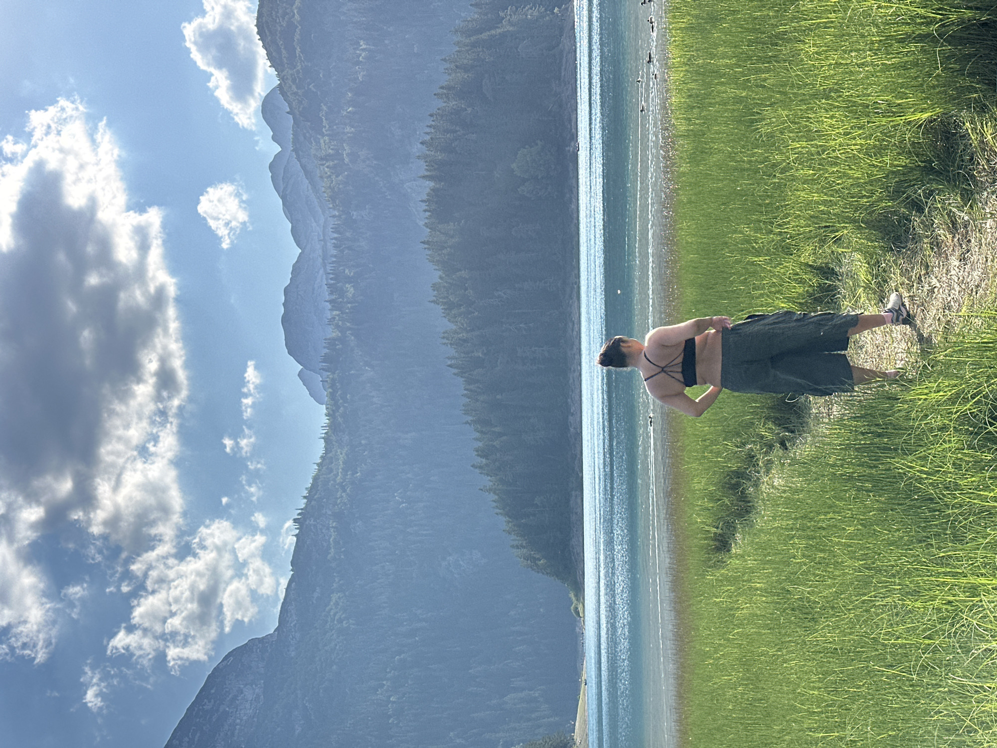 A woman with her back to the camera stands in a grassy field overlooking a large, blue lake. She is wearing a black sports bra and dark green shorts. The lake is surrounded by lush green forests and dramatic mountains, under a bright blue sky with scattered clouds. Ducks can be seen on the water in the distance. The location is identified as being in Pitomine, Montenegro.