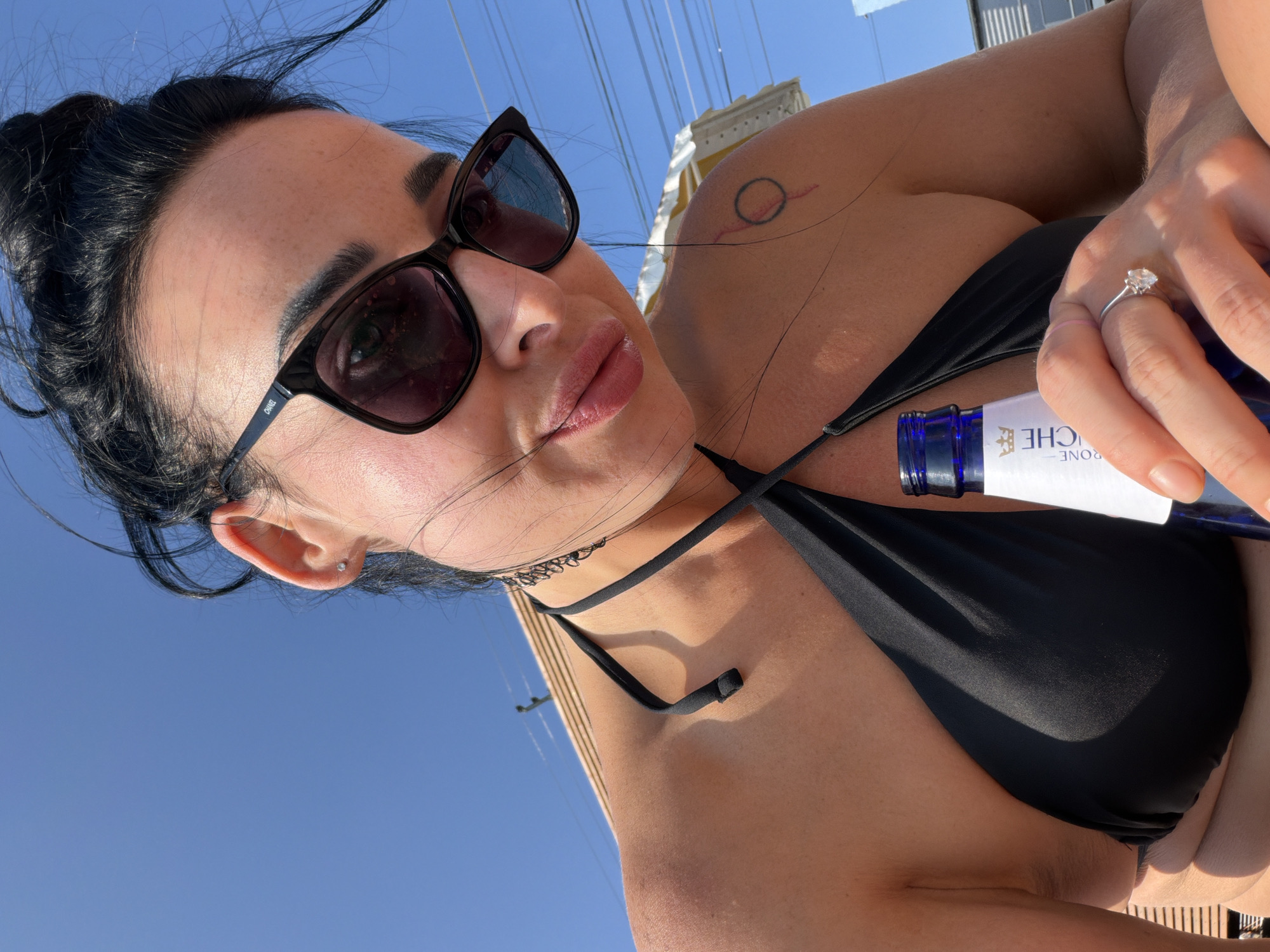 A close-up shot captures a woman from the chest up, looking towards the camera under a bright blue sky. She is wearing a black bikini top and dark sunglasses. Her dark hair is pulled up into a bun, with loose strands framing her face. A small, diamond stud earring is visible in her left ear. A tattoo of a circle with a line through it is on her right shoulder. In her right hand, she is holding a bottle with a blue cap, its label partially visible. A diamond ring is on her left ring finger. The background shows a glimpse of what appears to be a beach umbrella and some overhead wires. The lighting suggests it is daytime. The provided location information of Shepsi, Russia is not visually verifiable from the image content.