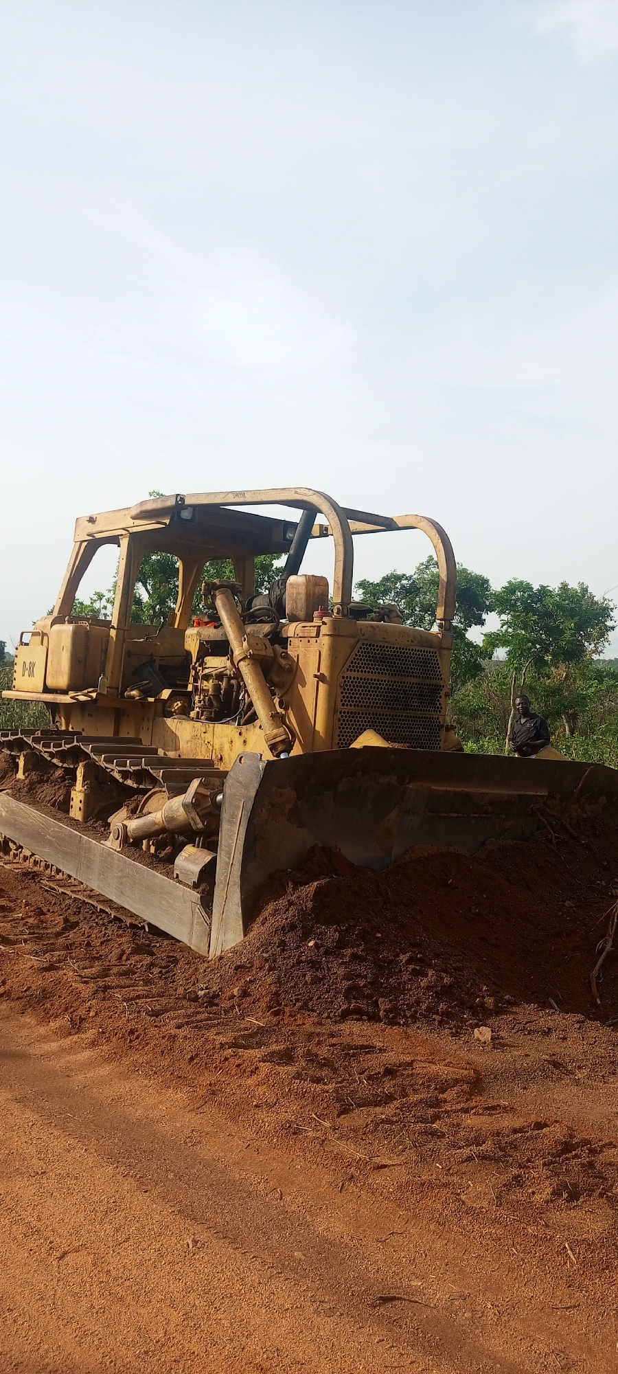 A yellow, weathered D-8K bulldozer is the prominent subject in this outdoor scene, set in Otunade, Nigeria. The heavy machinery, a Caterpillar D8K model indicated by the "D-8K" text visible on its side, occupies the foreground and midground. It is situated on and surrounded by reddish-brown earth, with its large front blade lowered and pushing against a substantial pile of the same soil, suggesting recent land-clearing or earthmoving activity. The bulldozer shows clear signs of heavy use, with dirt, rust, and wear across its metallic frame, tracks, and exposed mechanical parts. An open cab, protected by a robust rollover structure, is visible, and a plastic jerry can along with other items are placed on a platform near the cab.
In the middle-right background, a man stands partially obscured by the bulldozer's blade and surrounding foliage. He wears a dark shirt and appears to be observing the scene. The background is characterized by dense green trees and vegetation, indicative of a lush, possibly rural, landscape. The sky above is bright and appears overcast or hazy, diffusing the light evenly across the scene. The overall impression is that of a construction or development site in a tropical environment, captured during the daytime.