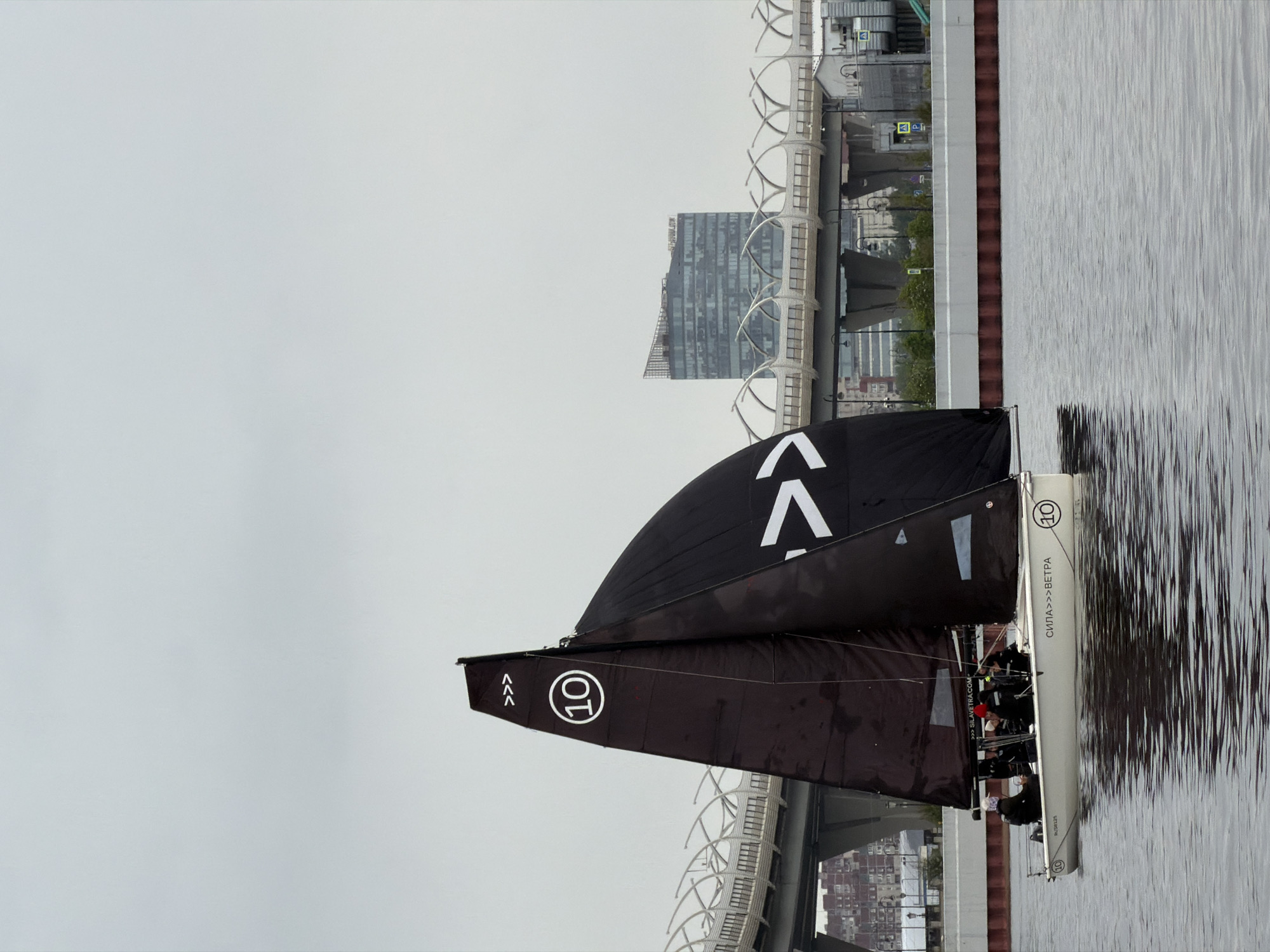 Captured from a low angle, the photo presents a modern sailboat navigating the waters near the urban landscape of St. Petersburg, Russia. The boat features prominent black sails adorned with white symbols and numbers, including a "10" and chevron patterns. The hull of the boat is white with black lettering and numbers, with "RUS8125" visible towards the front and ">SILAVETRA.COM", "СИЛА>>>ΒΕΤΡΑ" and "(10)" visible towards the back.
A group of individuals are visible onboard, engaged in sailing the vessel. The calm waters provide a clear reflection of the boat and its sails. In the background, modern architecture is visible, including a towering glass-clad building and elevated roadways with latticework structures. The sky is overcast, contributing to the muted lighting of the scene. The perspective emphasizes the boat's proximity to the cityscape.