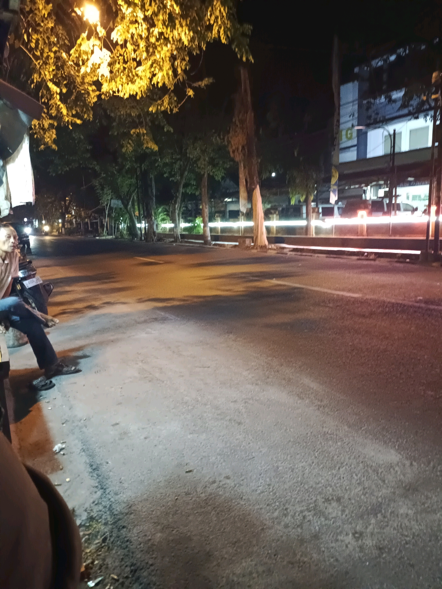 The image captures a nighttime scene on a street in Lamongan, Indonesia. The foreground shows a paved road with a white line marking. On the left side, a man is seated on what appears to be a motorcycle, leaning back. He is wearing a light-colored shirt and dark pants, with sandals on his feet. Adjacent to him is a structure, possibly a stall or shelter, with some white fabric hanging from it. The background reveals trees lining the street, illuminated by a bright yellow streetlamp overhead. Further down the road, buildings are visible, with some lights on. There are streaks of light in the distance, indicating moving vehicles. The overall atmosphere is that of a quiet street corner at night.