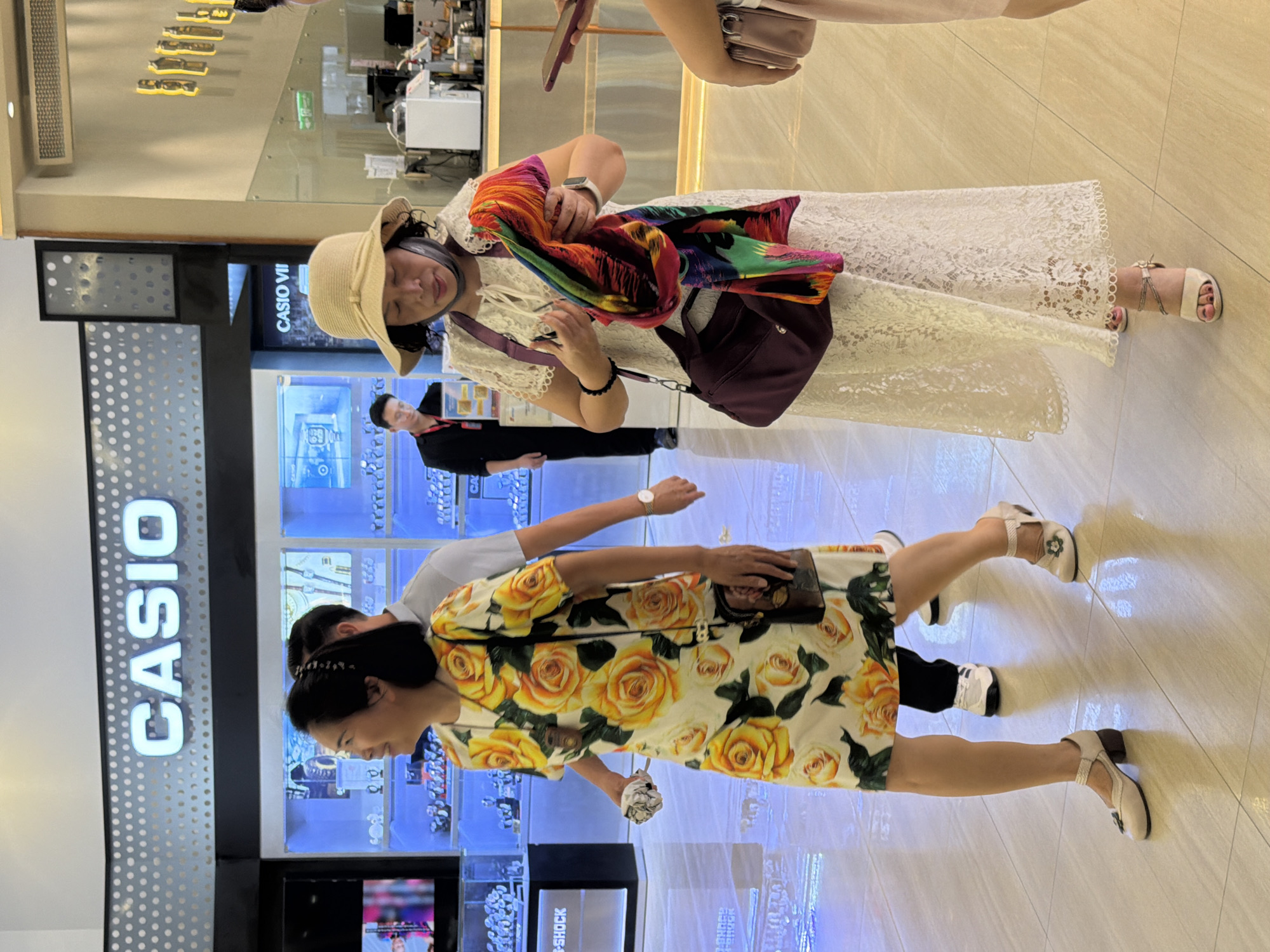 In Nha Trang, Vietnam, two women are walking through a brightly lit shopping mall, with a prominent "CASIO" sign visible in the background. The woman on the left, wearing a vibrant yellow dress with a rose print, is looking to her left with a slight smile. She is carrying a small object in her right hand. The woman on the right, wearing a white lace dress and a straw hat, is holding a colorful scarf and looking down at it. She has a purple shoulder bag slung across her body. A man in a black suit is visible in the background, standing near a display of watches. The floor is tiled with a light-colored, reflective material.