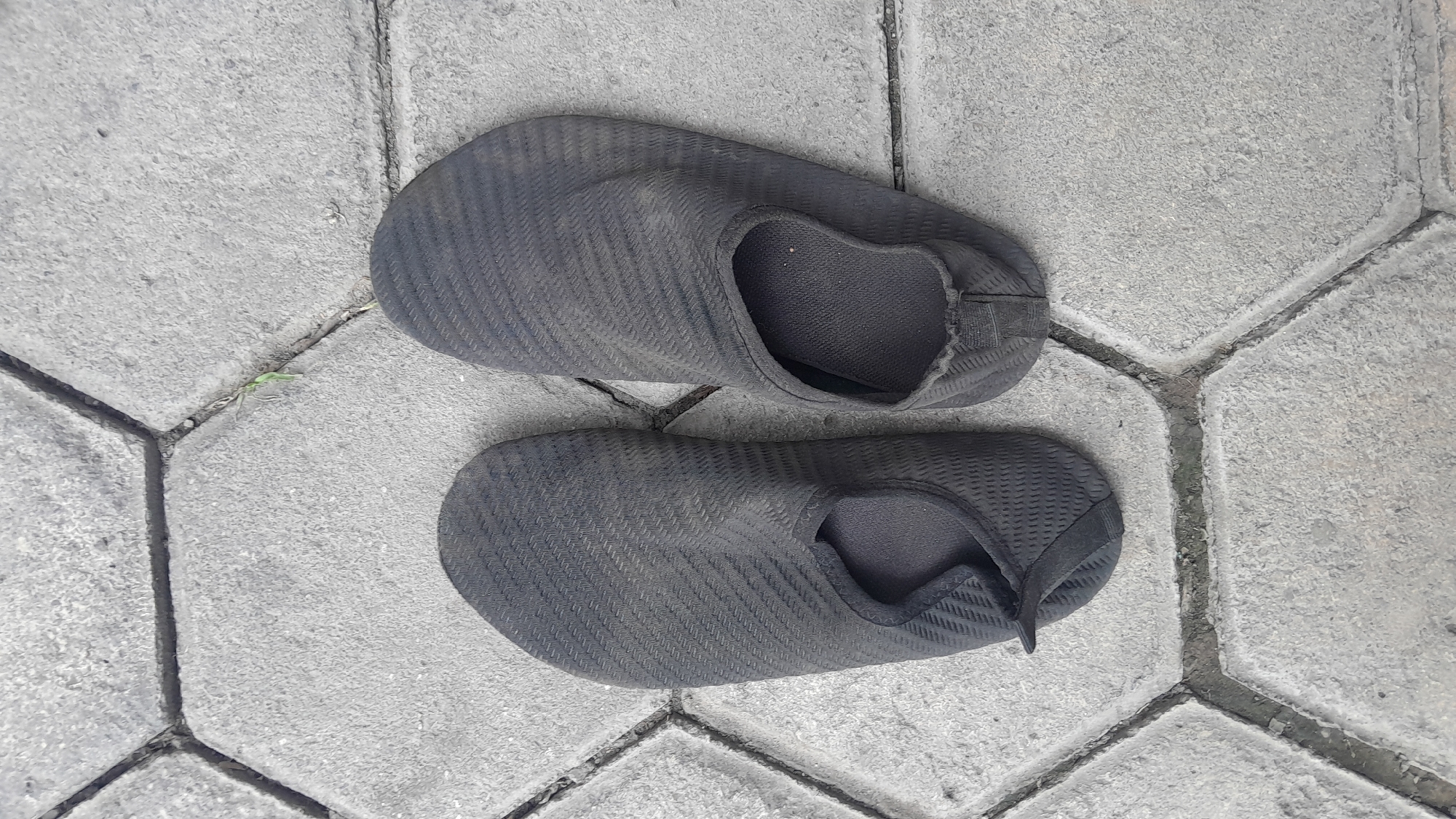 A pair of dark grey water shoes are placed on a pavement made of hexagonal concrete blocks. The shoes are oriented side-by-side, with the left shoe slightly angled towards the viewer and the right shoe positioned more directly facing upwards. Both shoes appear to be made of a flexible, mesh-like material with a textured pattern. They are slightly dusty, indicating they have been used. The pavement is rough and appears weathered, with visible gaps between the blocks filled with dirt and some small green weeds growing in one of the gaps. The scene is captured from a top-down perspective. The location is identified as Sambong, Indonesia.