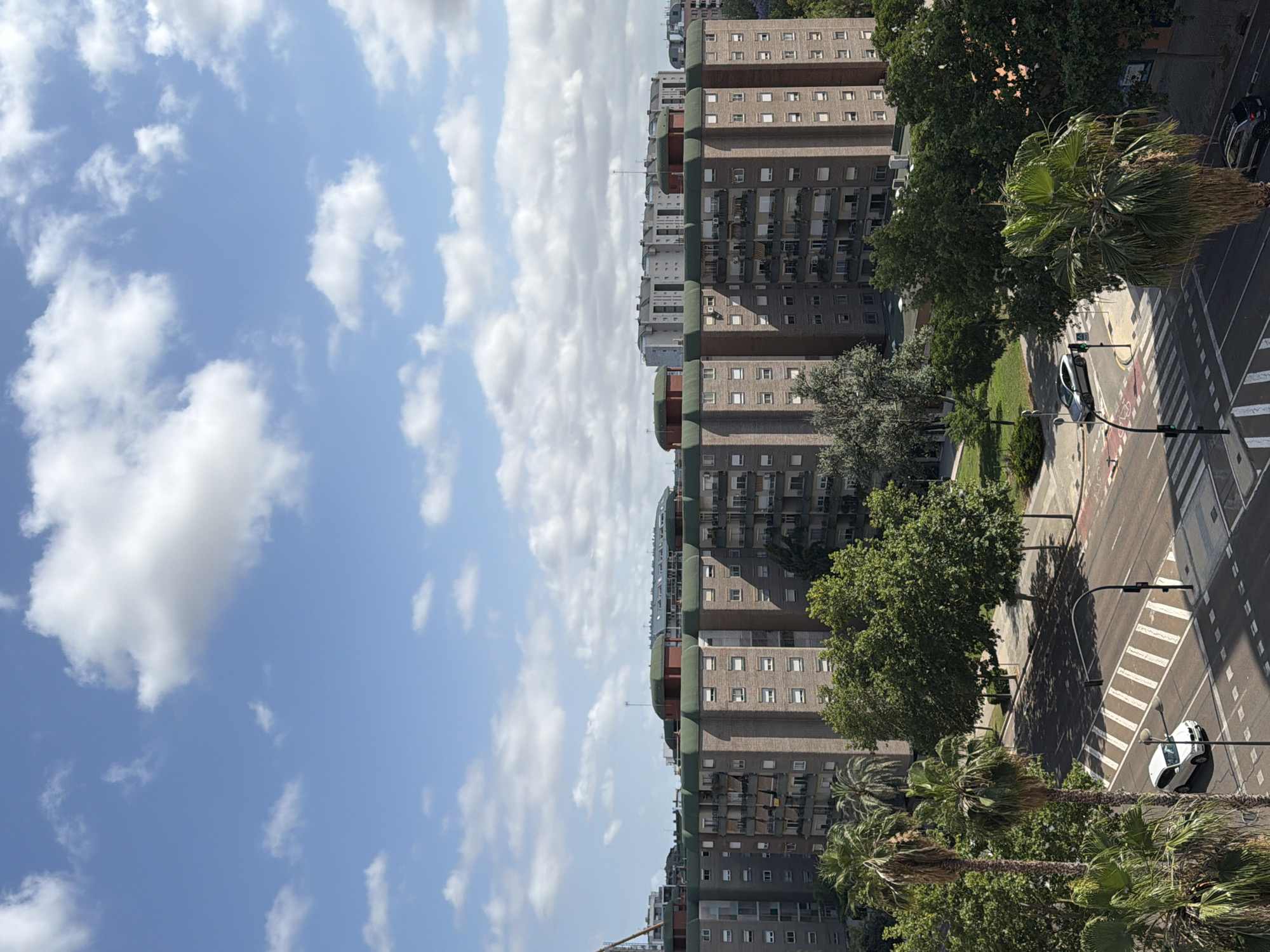 This is a high-angle shot of a city street with apartment buildings and trees. In the foreground, there are several palm trees and lush green trees. A white car is visible on the street, stopped at a crosswalk. Across the street, there is another car and a dark-colored vehicle parked. The background features a row of tall, brick apartment buildings with green roofs, and further in the distance, more buildings are visible. The sky is blue with scattered white clouds.