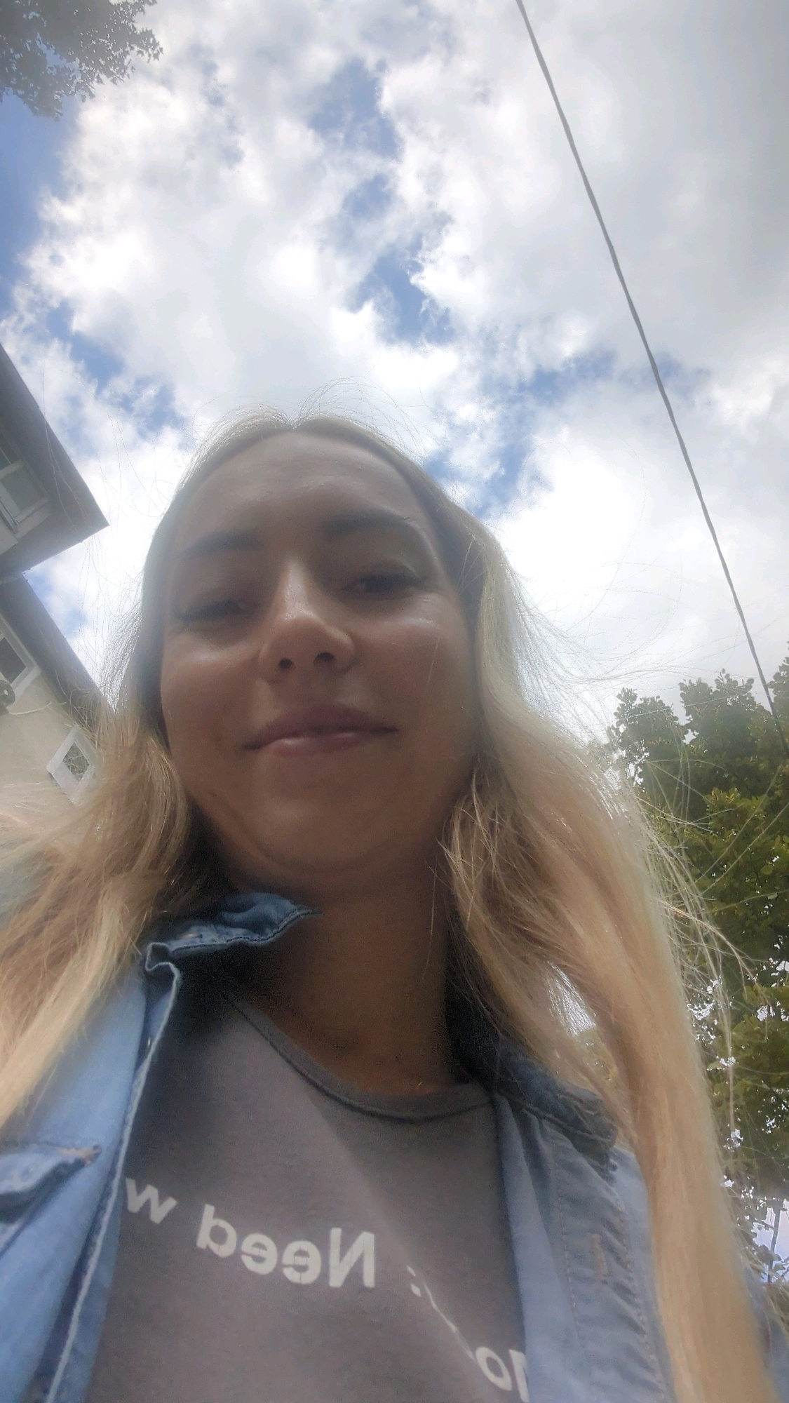 A low-angle, medium shot captures a young woman from the chest up, with her face close to the camera. She has long, blonde hair that falls over her shoulders. She is wearing a grey t-shirt with white lettering that reads "w been: or" and a blue denim jacket over it. The background consists of a cloudy sky, with some trees and a building visible to the left. A power line runs diagonally through the upper right portion of the frame. The image was taken outdoors, likely in Chișinău, Moldova, given the architectural style of the building and the presence of power lines. The scene is neutral, with no discernible action or interaction taking place.