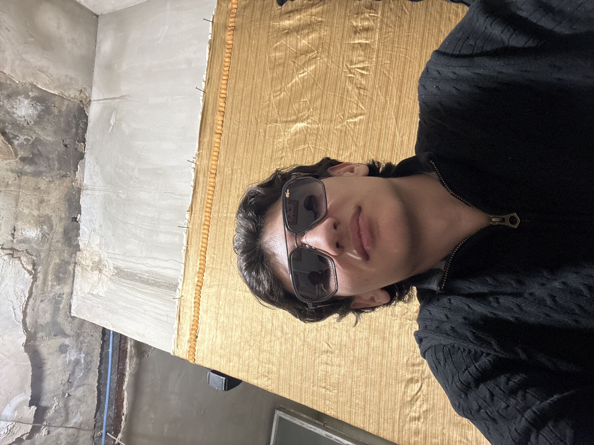 A young person, likely male, is captured in a close-up, chest-up selfie, situated indoors in Yerevan, Armenia. The subject has dark, wavy hair and is wearing dark, rectangular sunglasses, which prominently display a small Lacoste crocodile logo on the right lens. Their expression is neutral or slightly contemplative. They are dressed in a dark, possibly navy or black, cable-knit sweater with a brass-colored zipper at the collar.
The background is split between a textured golden fabric and an unfinished or aged wall. To the subject's right, a large section of gold-colored, vertically pleated fabric, resembling a curtain or tapestry, is visible, secured by several nails along its edge. To their left, the wall shows exposed concrete and plaster, with visible cracks and underlying building materials, suggesting a state of renovation or an older structure. A blue cable runs vertically along this raw wall segment. Below the golden fabric, a dark surface with a small, rectangular reflection or window is partially seen. The lighting appears to be artificial. The activity is a self-portrait, with the person posing for the camera.